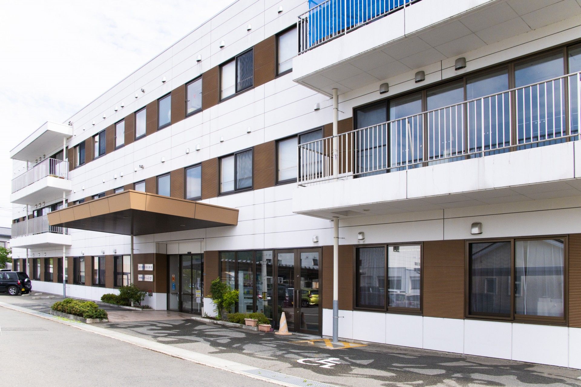 白色の外壁の建物で、玄関前に大きな屋根の備わった車寄せがある。玄関周りには木が植えられ、広い駐車場が整備されている。