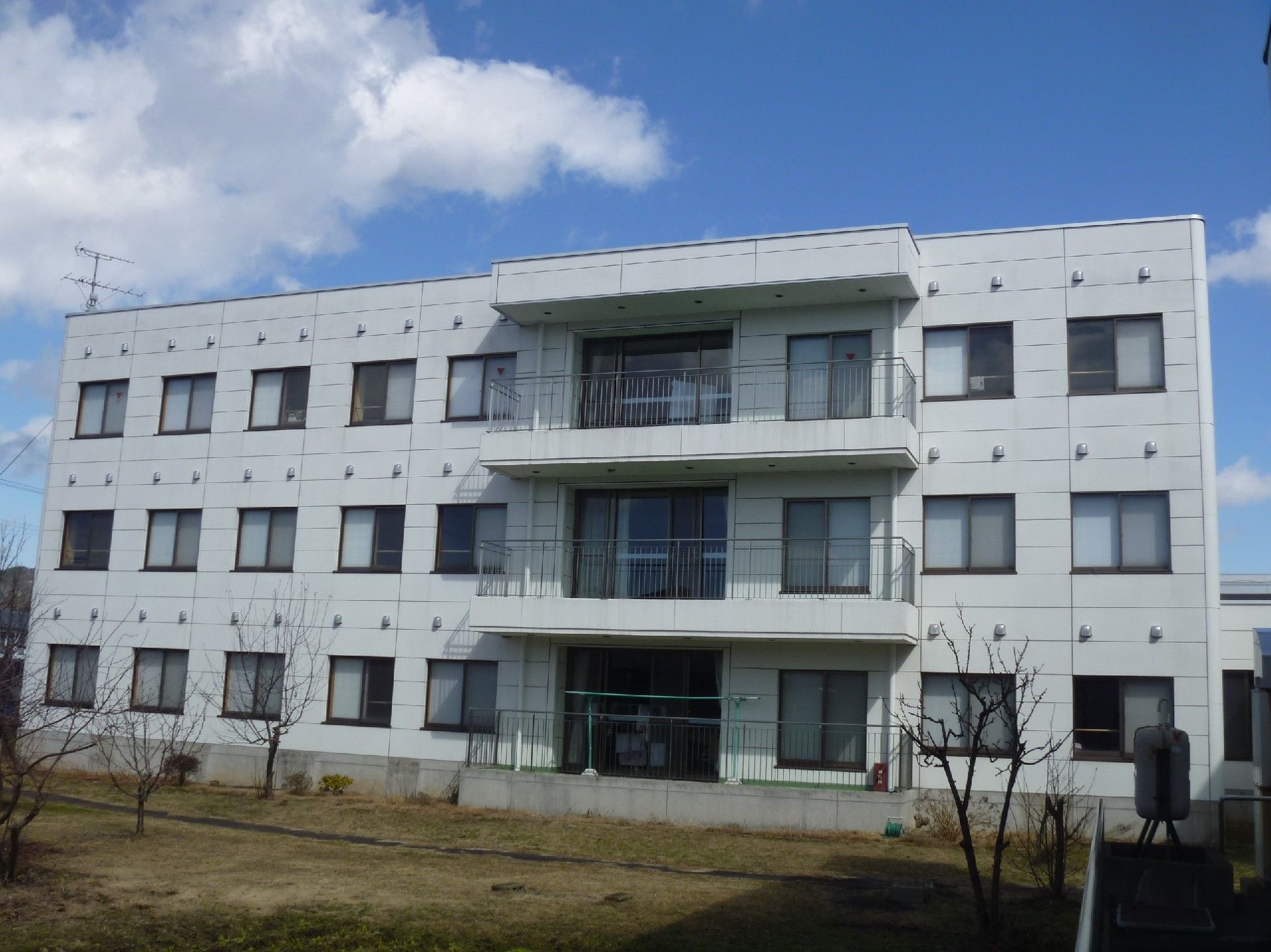 施設は白い３階建ての建物である。各階に転落防止柵付きのバルコニーが設けられており、１階には物干し台が設置されている。