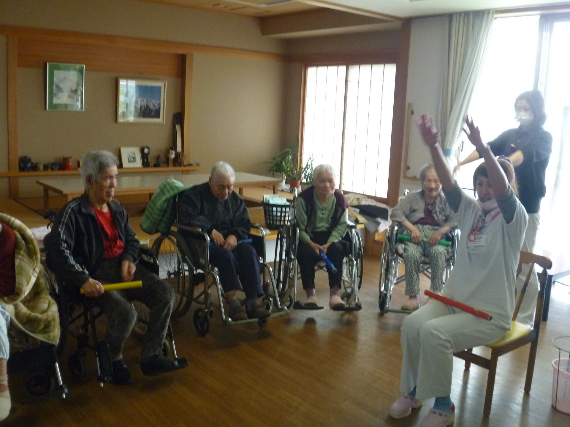 車椅子をご利用の方も広い部屋に集まって、指導員さんの指導のもと、バトンのような物を使った体操をしている。