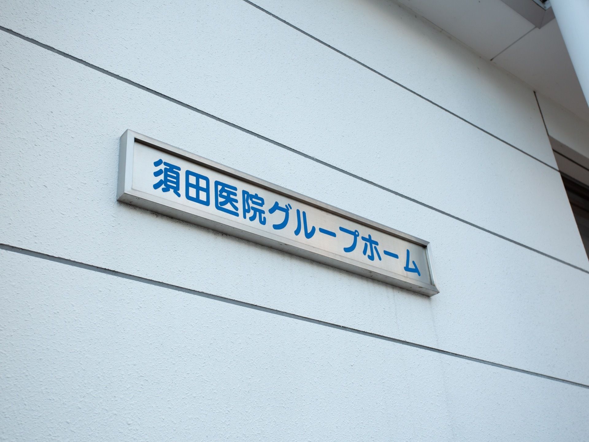 白色の施設の建物の外壁に、施設名が記された看板が取り付けられている。施設名はシルバーのプレートに青色の文字で書かれている。