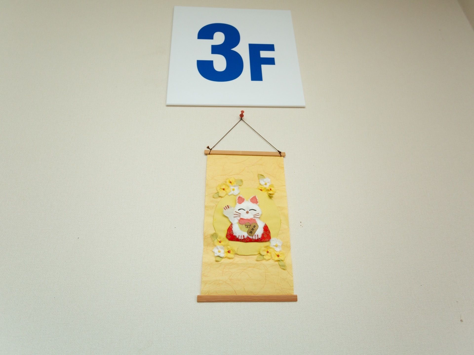 白色の壁に3Fを示す案内板が備わり、その下には黄色を基調としたタペストリーが飾られている。タペストリーの中央には招き猫がある。