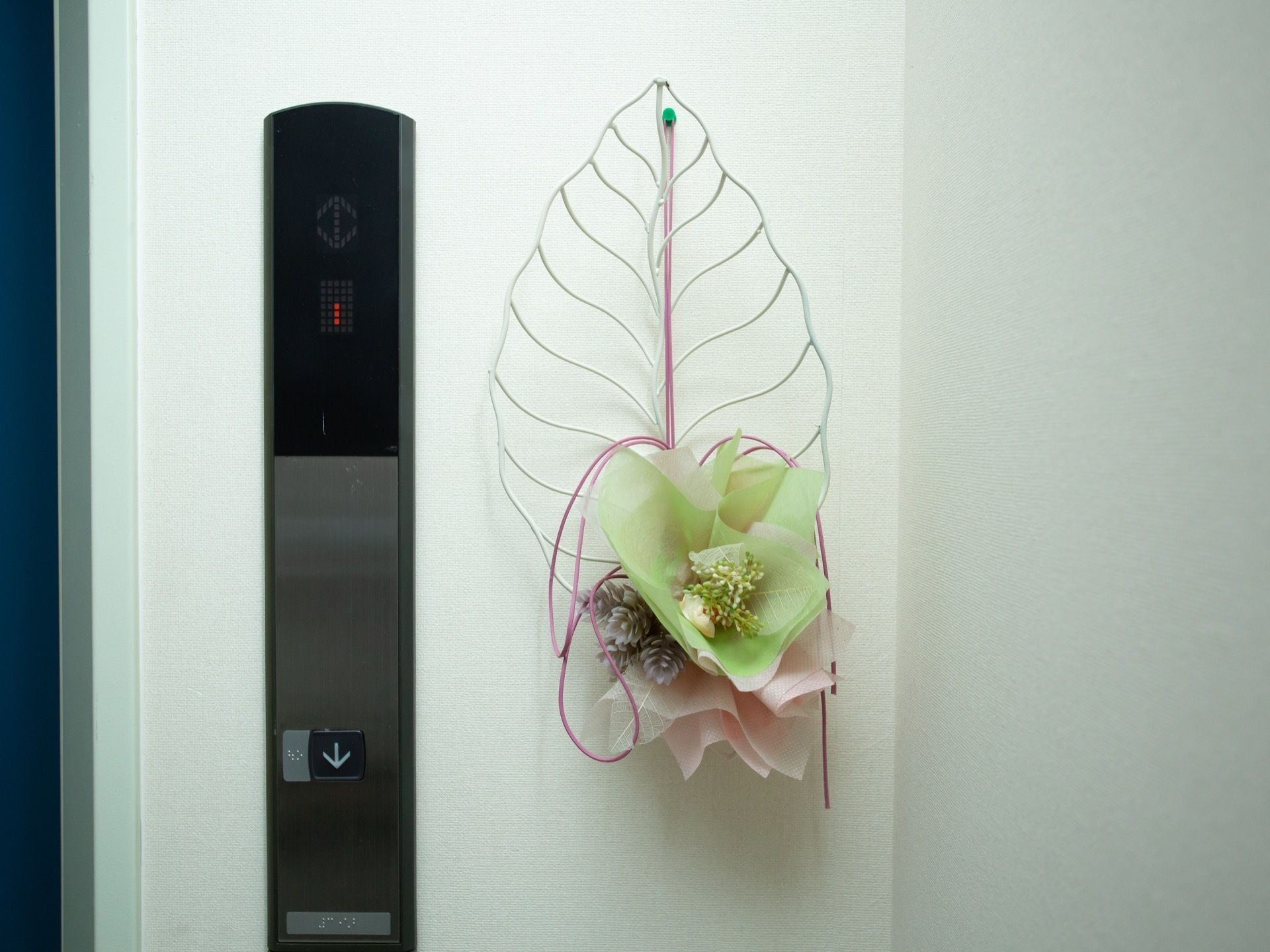 エレベーターの操作盤の横に飾られたインテリアである。白色のワイヤーで葉っぱを形作り、淡い緑とピンク色の花が添えられている。