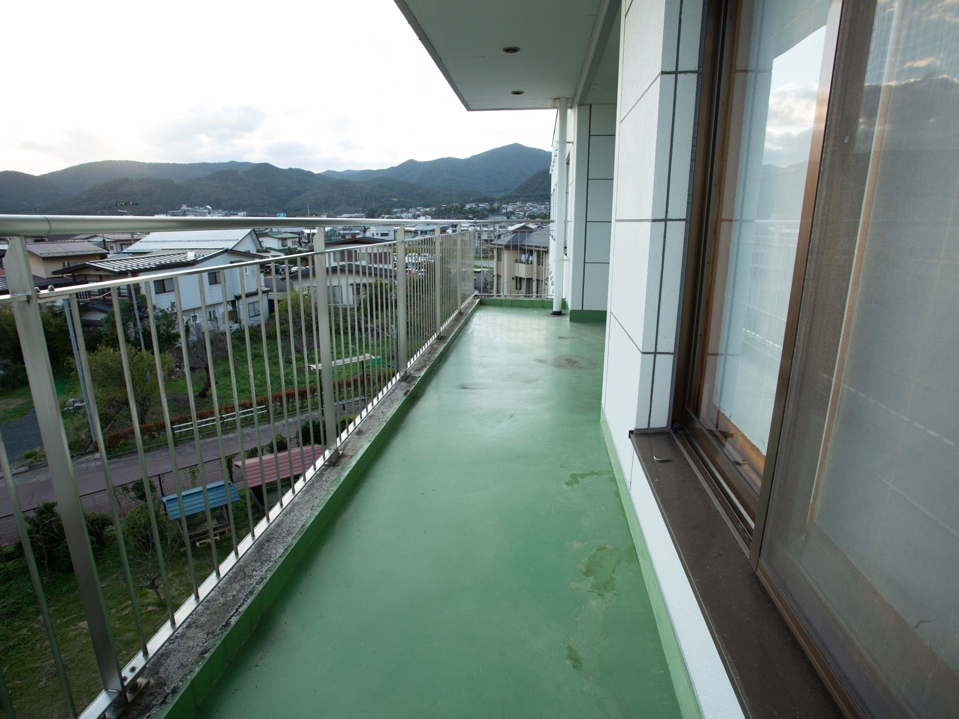 落ち着きのある緑色の床面のベランダである。施設の周辺には高い建物がなく、遠方に連なる山々を眺めることができる。