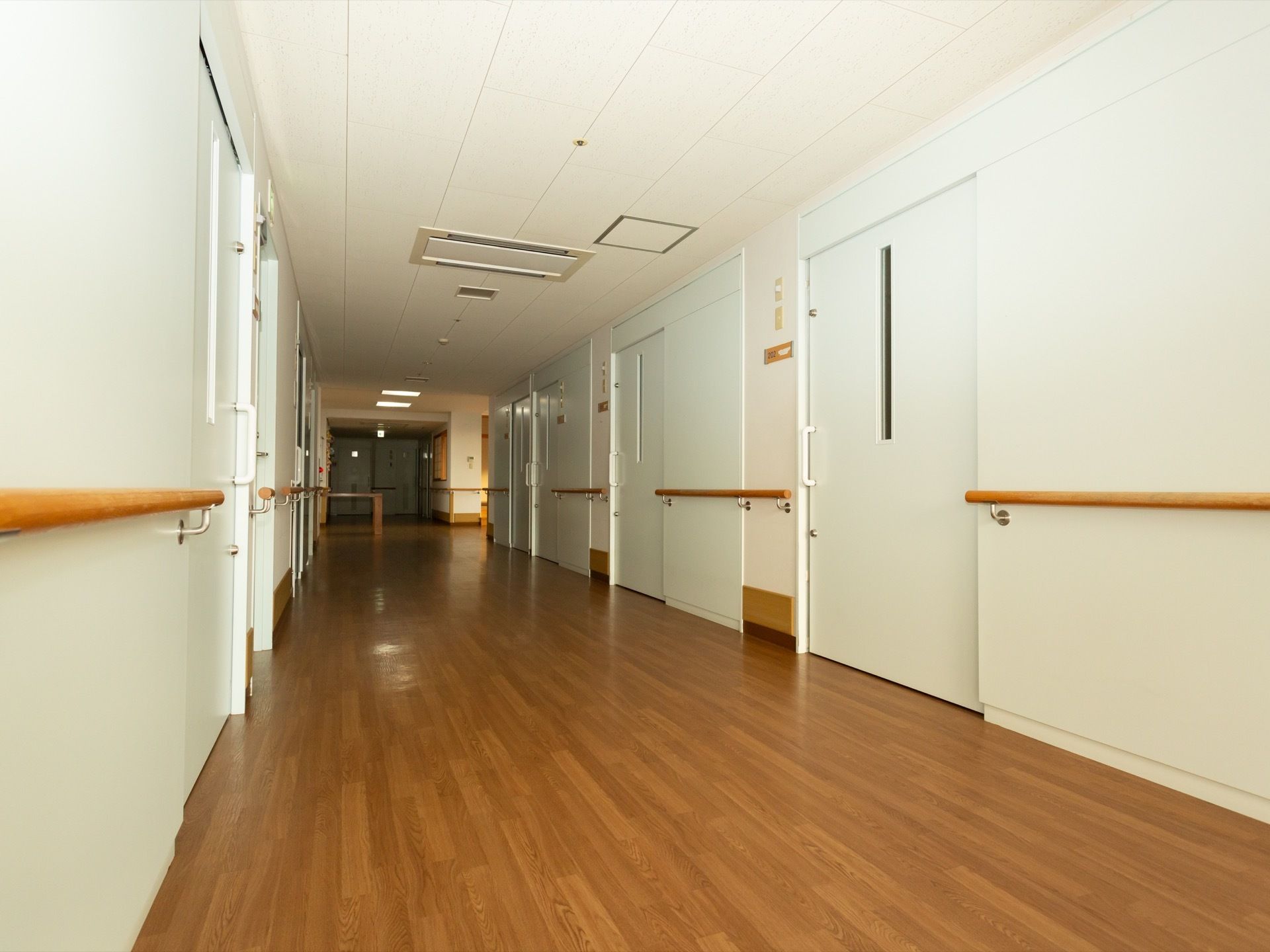 淡い茶色の木目のフローリングの通路である。通路の両側には、白色のスライド式のドアが並び、壁には手すりがある。