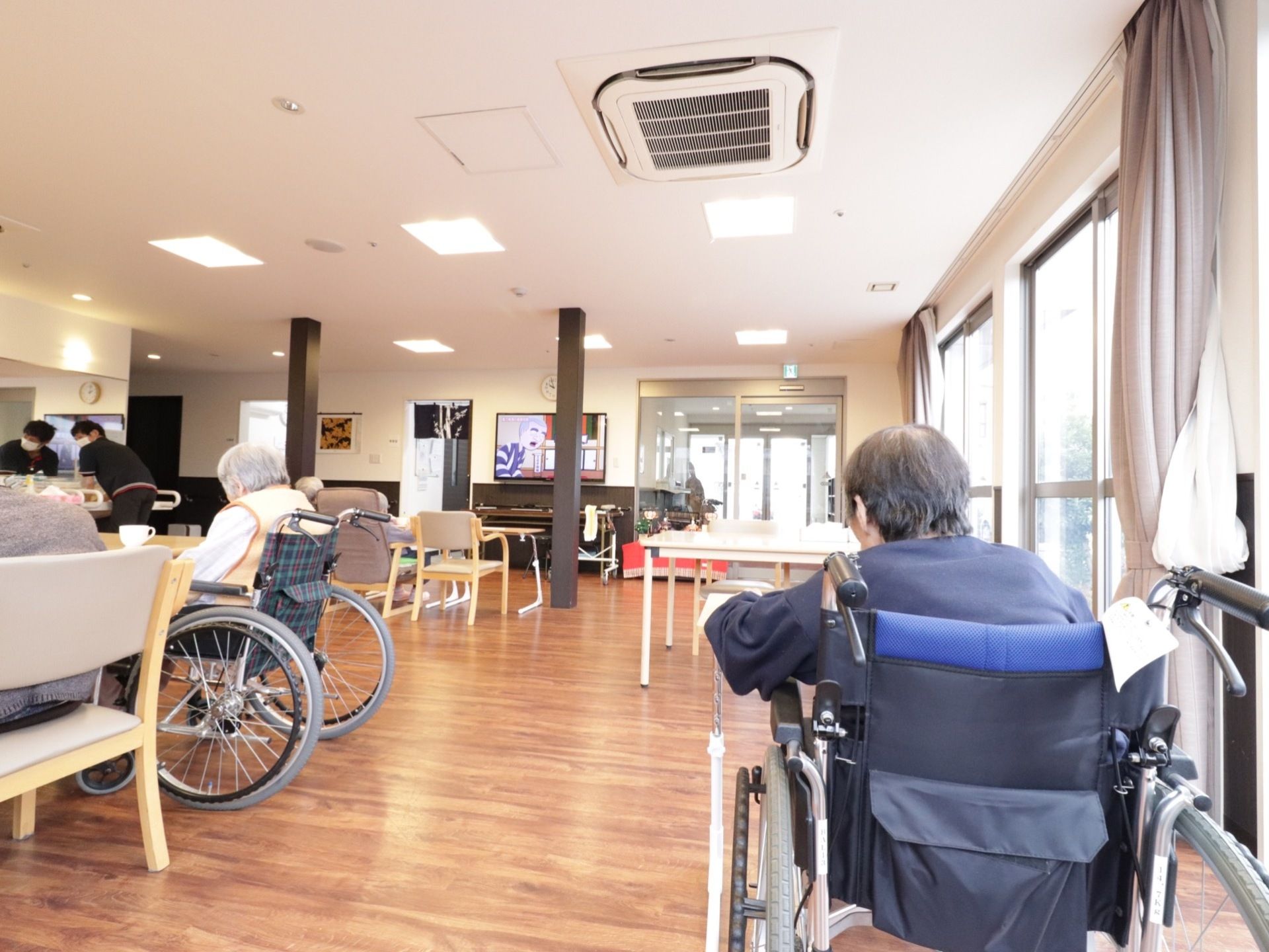 エントランスホールのすぐ側にある食堂は、外からの光がサンサンと差し込み大変明るくなっている。入居者様が座ってテレビを見ている。