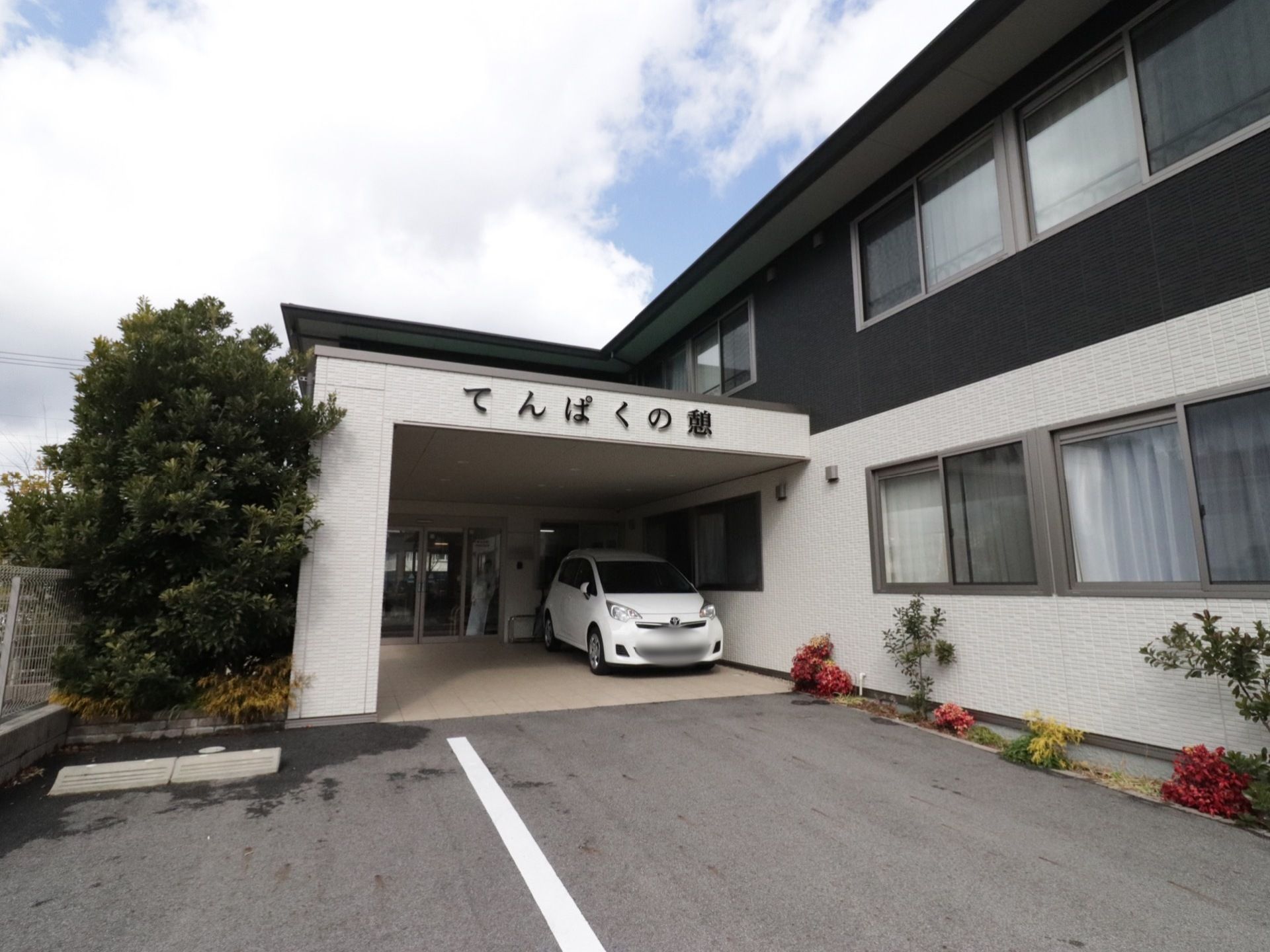 玄関前方の駐車スペースから、建物の外観を写したものである。右端には細長い植込みがあり、さまざまな草木が植えてある。