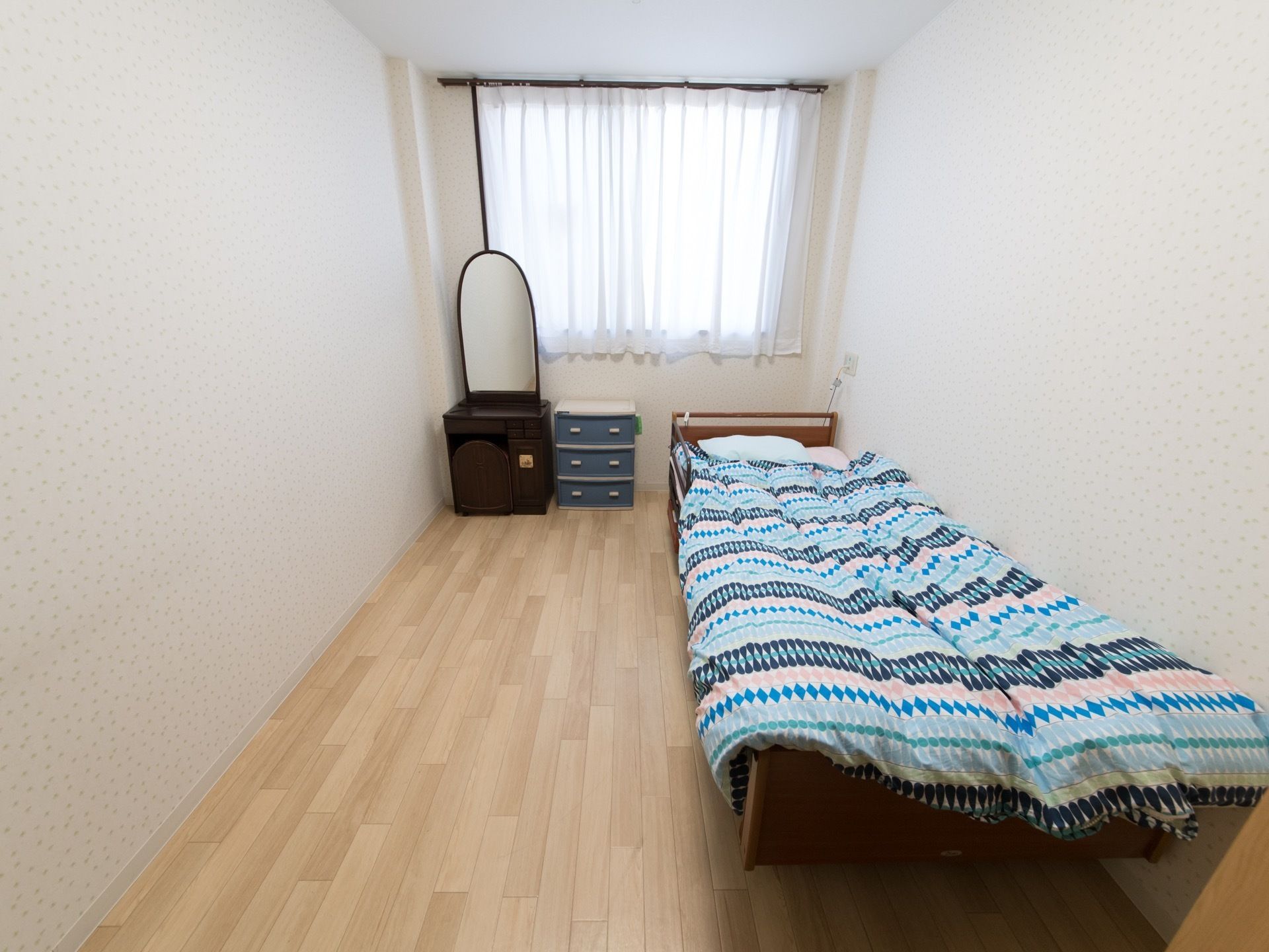 居室の一例。部屋の奥にレースカーテン付きの窓がある。右側にベッドが置かれ、左側に鏡台とタンスが置かれている。