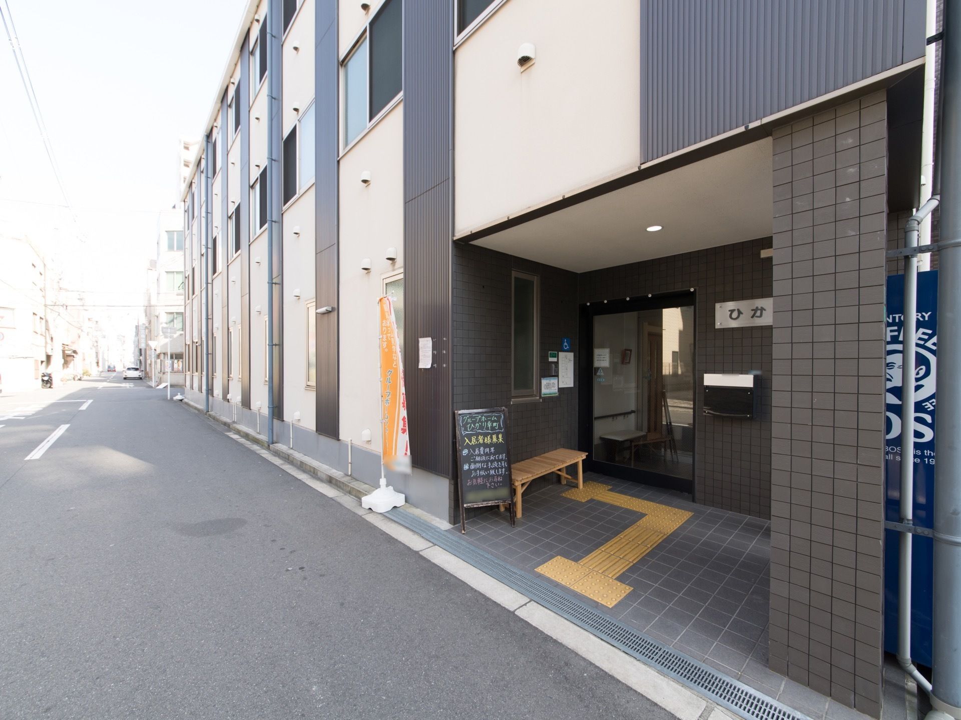 ベージュ色と茶色が組み合わされた落ち着いたデザインの外観である。玄関ポーチの右端には、自動販売機が設置されている。