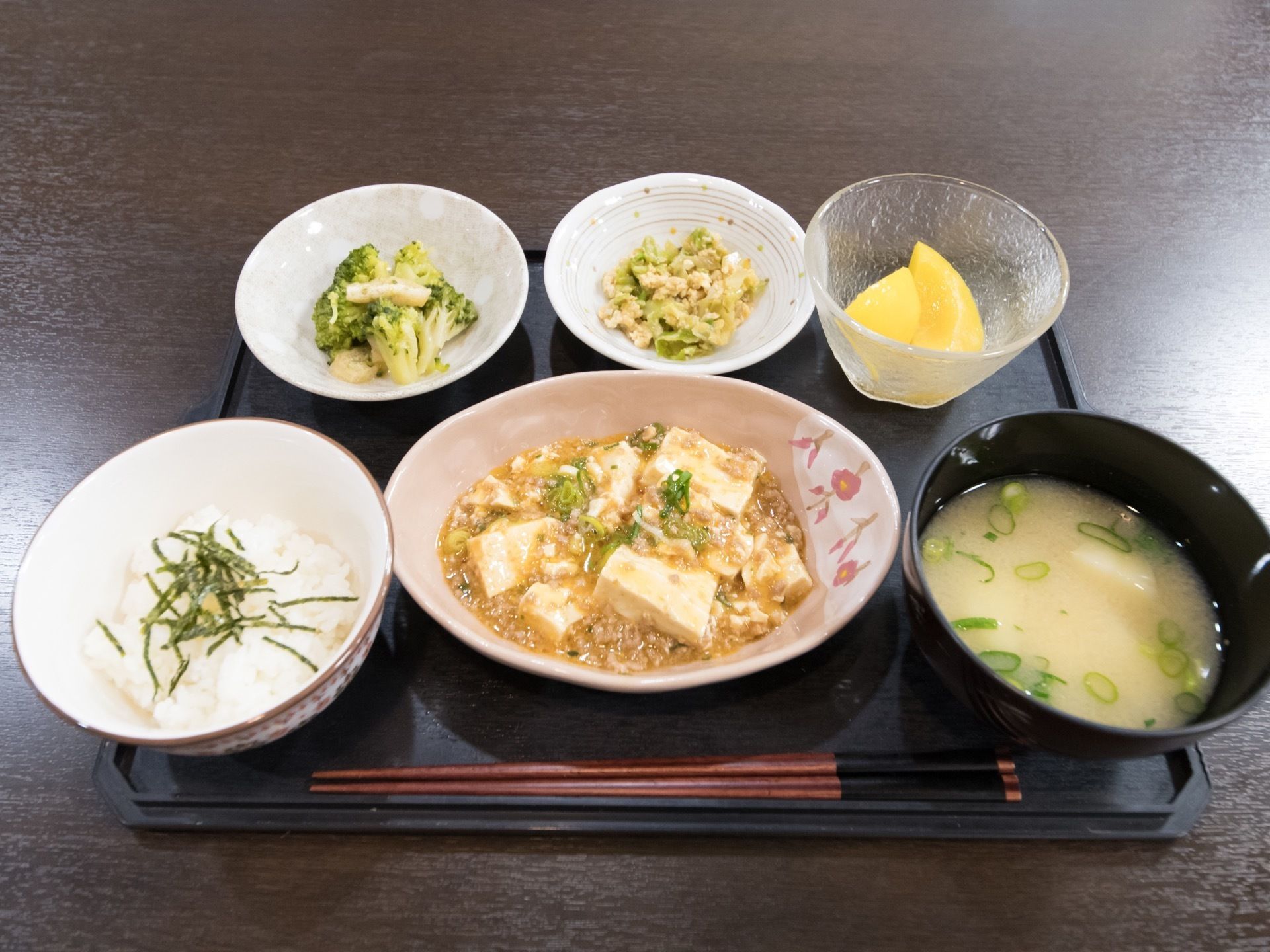 黒いトレーの上に、白米とお味噌汁、挽き肉とネギがたっぷり入った麻婆豆腐、小鉢に入ったブロッコリー、黄桃などが並んでいる。