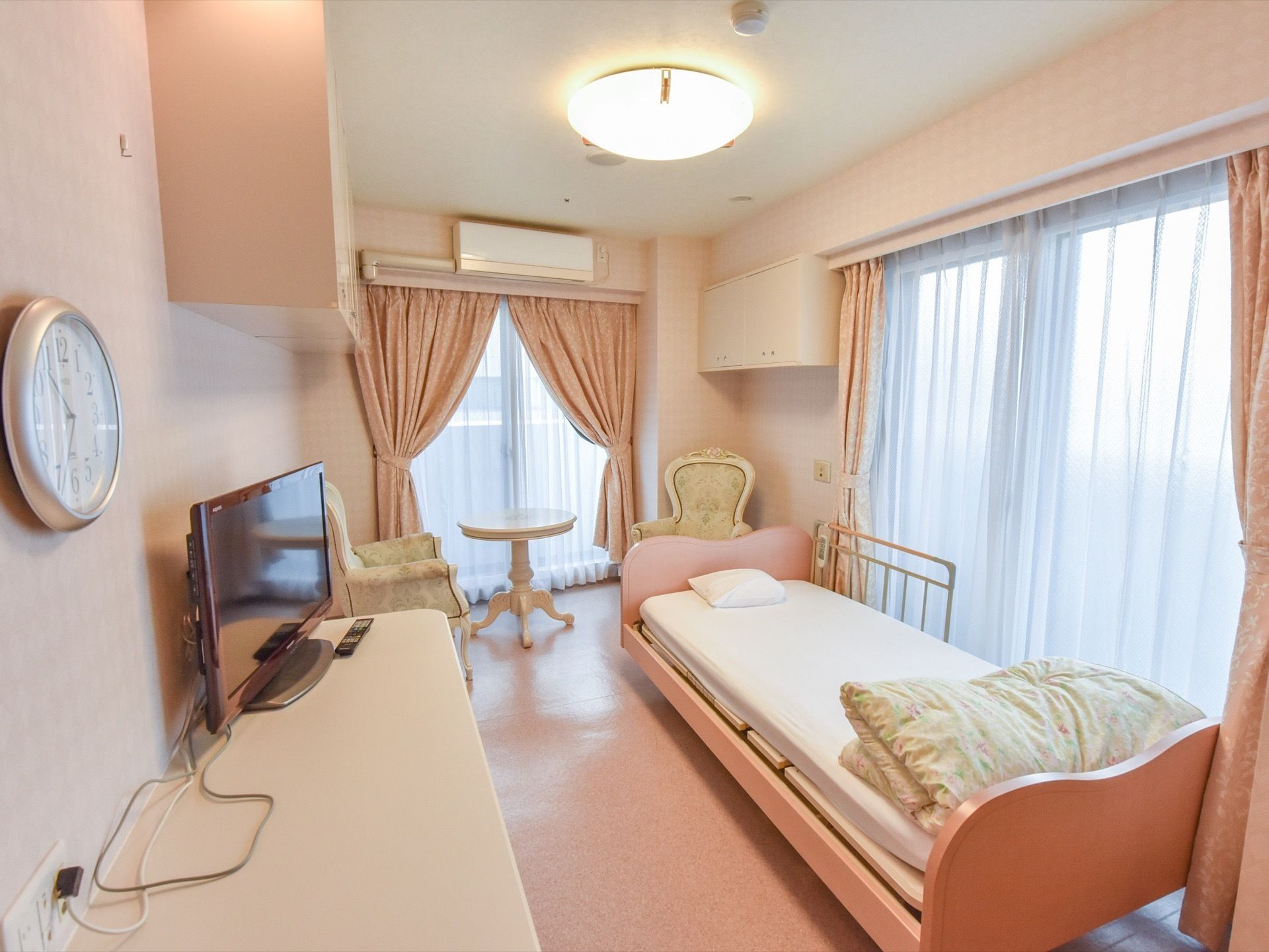ピンクとアイボリーを内装に採用しており、カーテンとチェアは上品な柄がついている。その他、部屋にはテレビとエアコン、備え付けの棚がある。