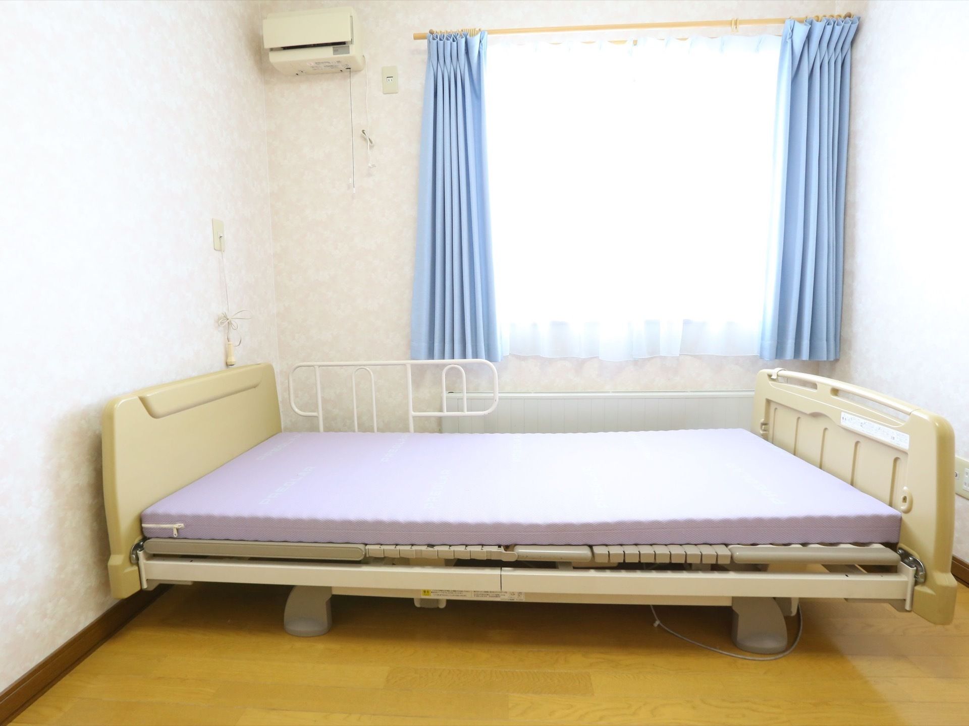 窓側の壁に電動ベッドが1台置かれており、厚めのマットが敷かれている。窓には白いレースと青いカーテンが掛けられている。