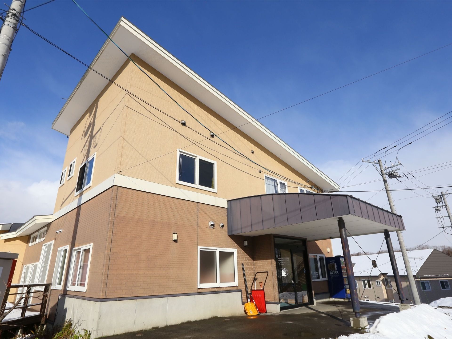 1階が茶色、2階がベージュ色の外壁の建物で、施設前は雪かきが行われている。周囲には住宅が立ち並んでいる。