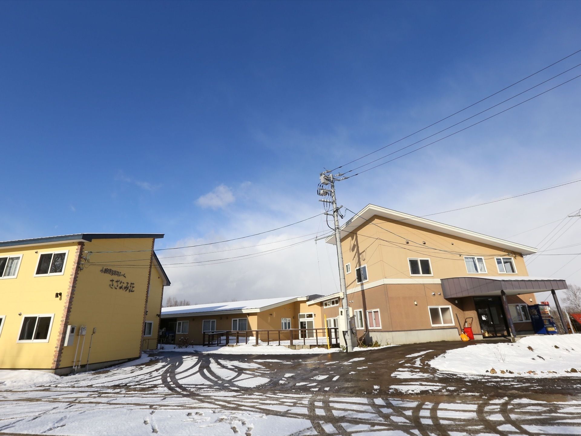 ベージュ調の2階建ての建物と平屋の建物になっている。自動販売機が設置されており、雪かき用のスコップが置かれている。