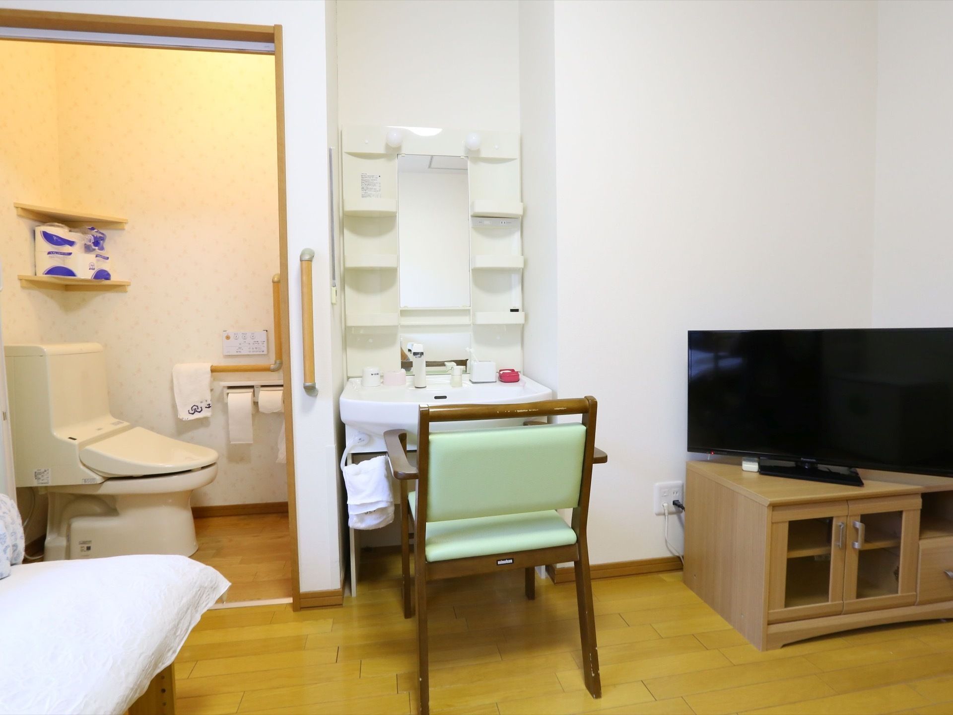 居室のトイレの扉は開かれている。設置された洗面化粧台の前には椅子が置かれ、チェストの上にはテレビが乗せられている。