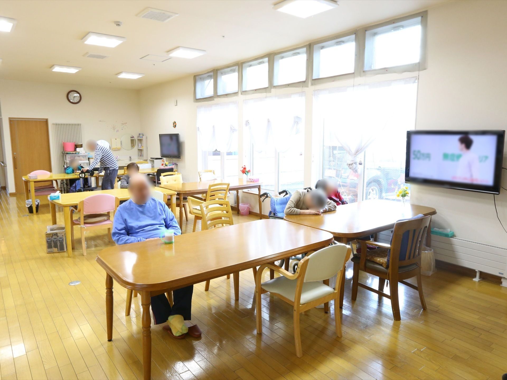 木目調のテーブルと椅子が配置された共有スペースである。テレビが点けられ、数名の入居者様の姿が見える。