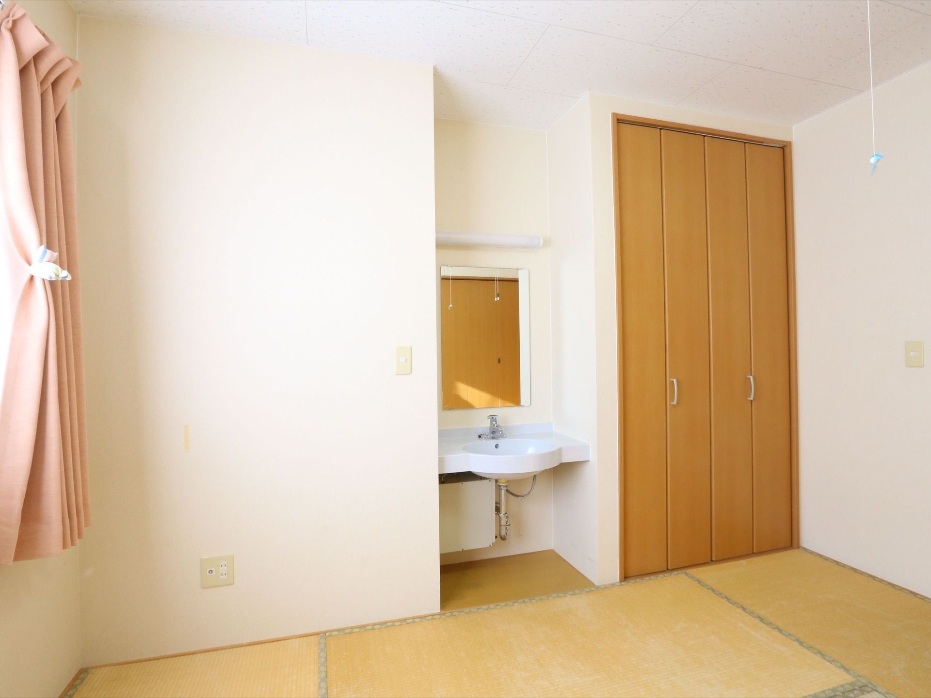 白い壁と畳敷きの入居者様の居室。窓にはピンクのカーテンが掛けられている。洗面台と鏡があり、ブラウンの扉のクローゼットがある。