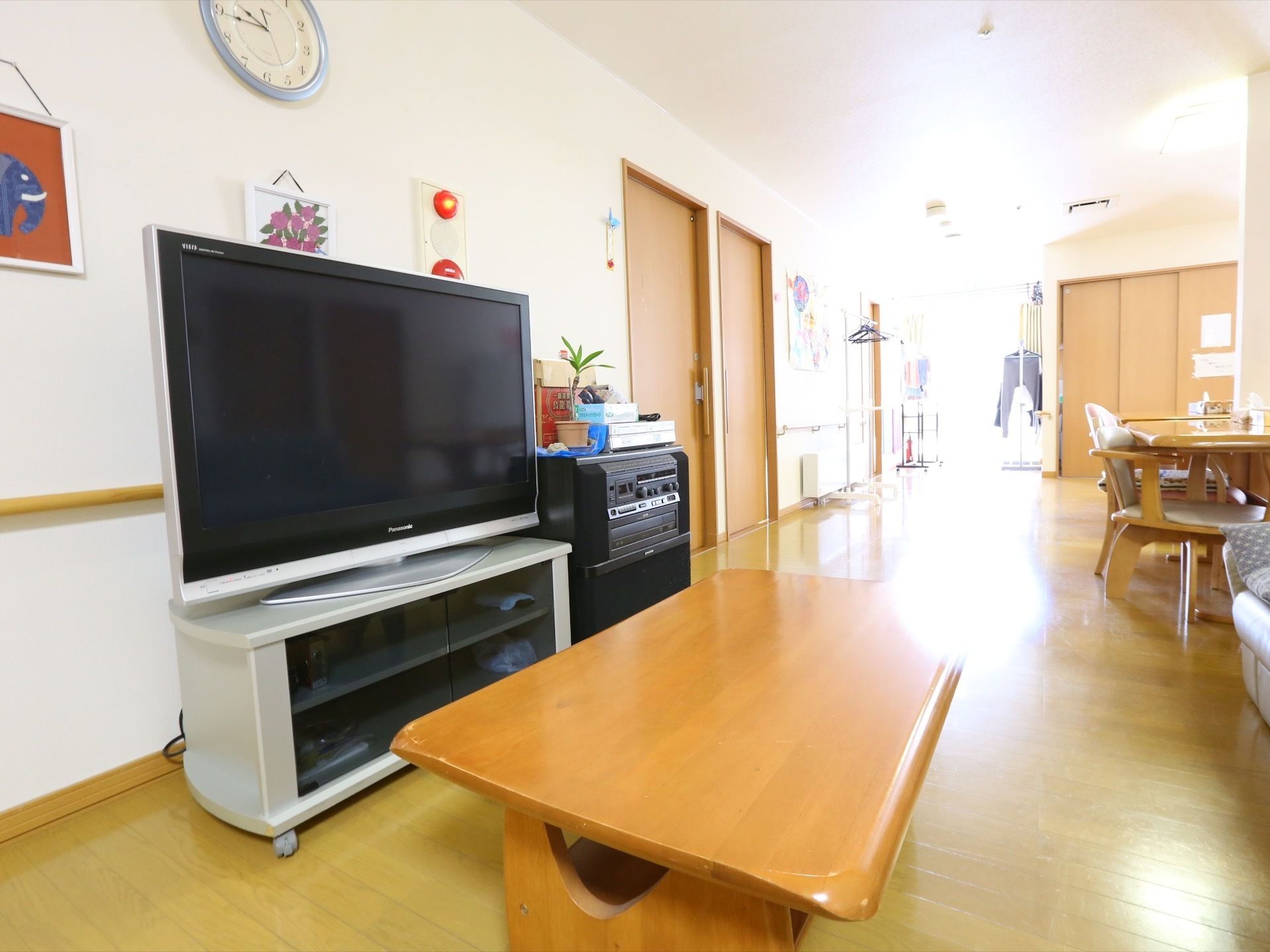 テーブルと椅子が並ぶ談話室の一角に、テレビ、ローテーブル、ソファが置かれている。壁には絵や時計が掛かっている。