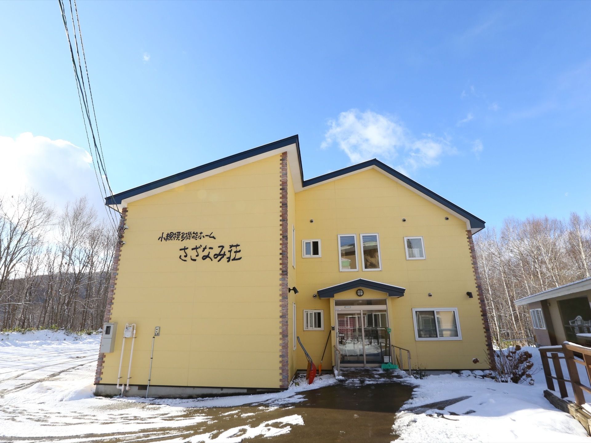 黄色い壁と斜めの屋根のデザインが特徴的な施設の外観。建物の裏には山と雑木林が茂っており、周囲には雪が積もっている。