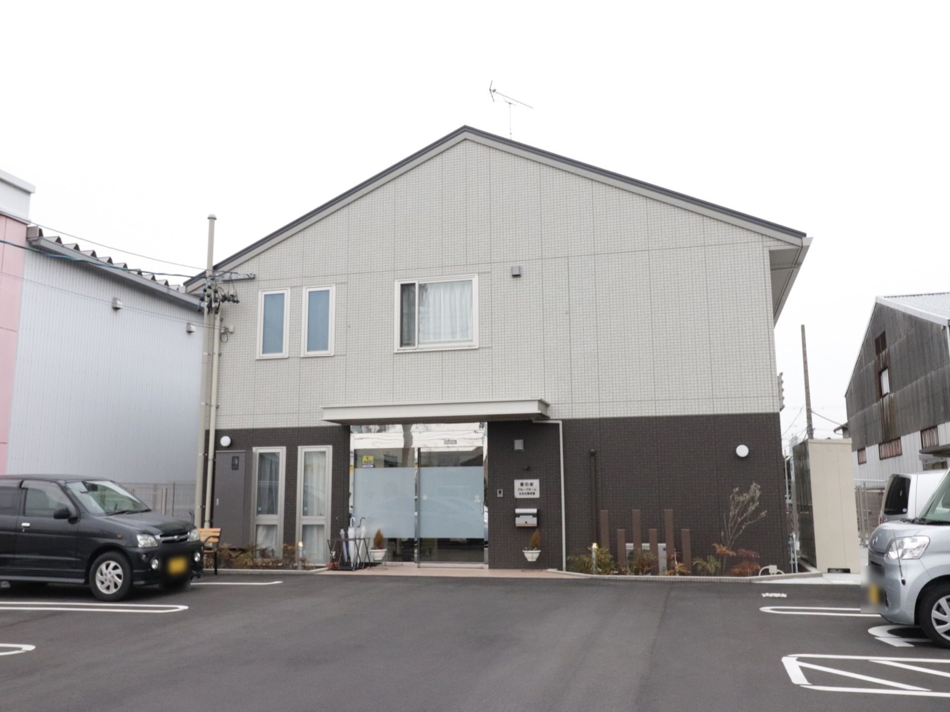 施設建物の外観。正面玄関の左右には植え込みが設けられている。敷地はアスファルト舗装の駐車場になっている。