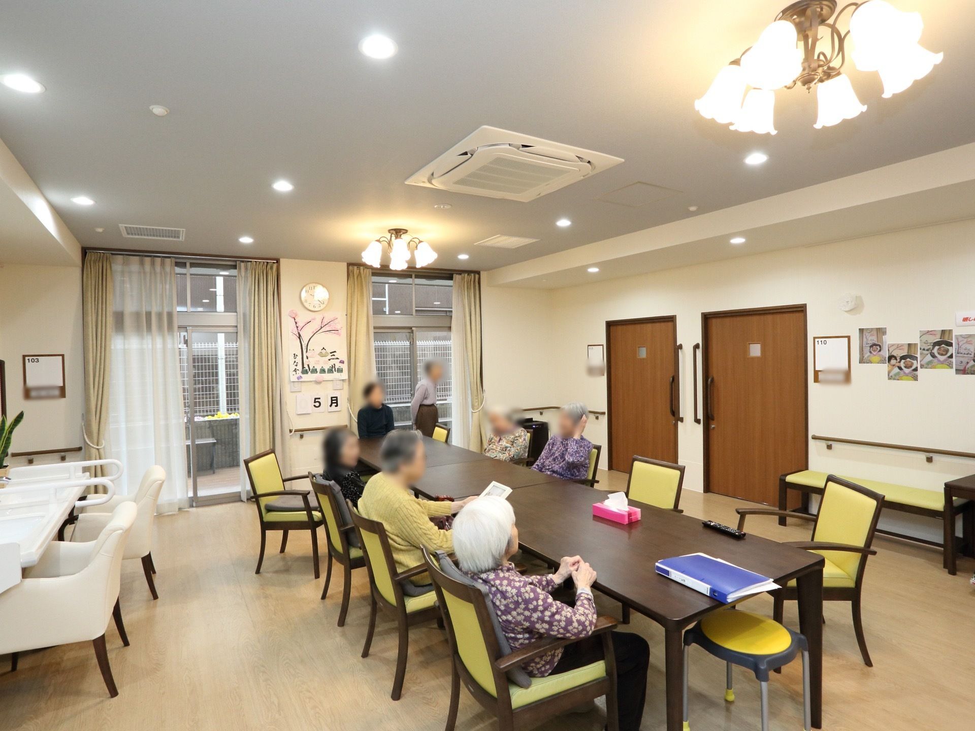 窓があり、カーテンが二重にかかっている。テーブルと肘掛け付きの椅子が設置してある。入居者様がくつろいでいる様子が写っている。