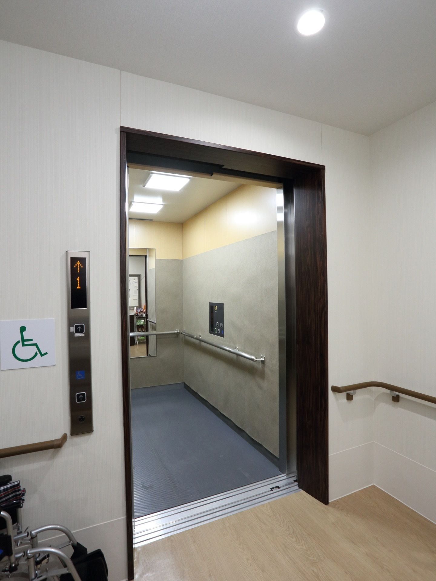 エレベーターの中に鏡と手すりが設置されている。天井に照明がついている。入口の左側の壁に車椅子が立てかけられている。