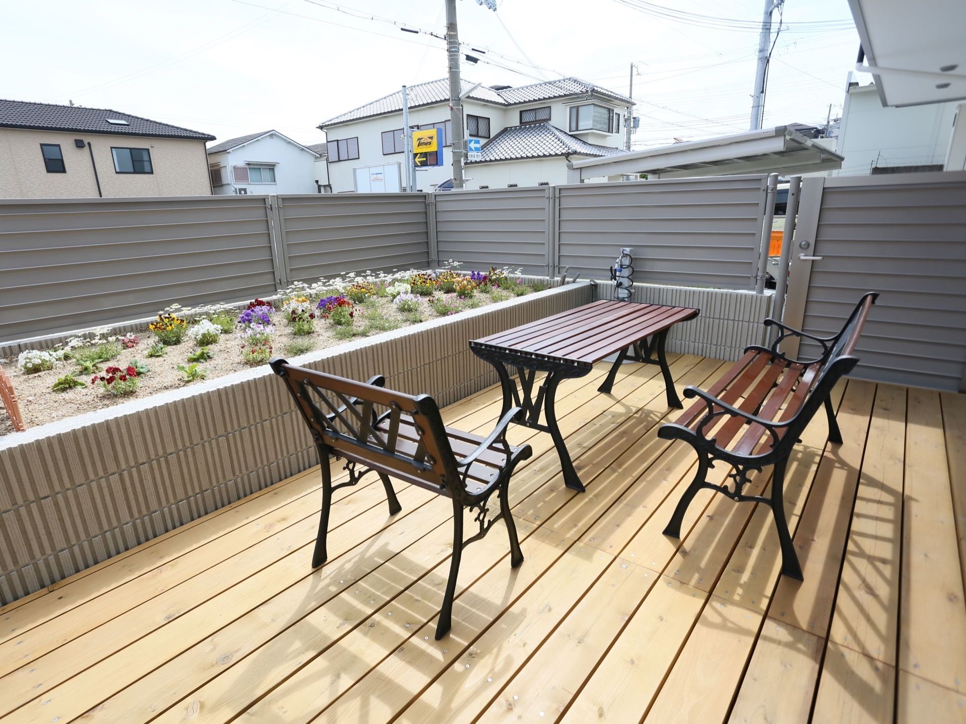 部屋の外に備えたバルコニーである。フェンス際に花壇を設け、木製の床の上にお洒落なデザインのテーブルと椅子を置いている。