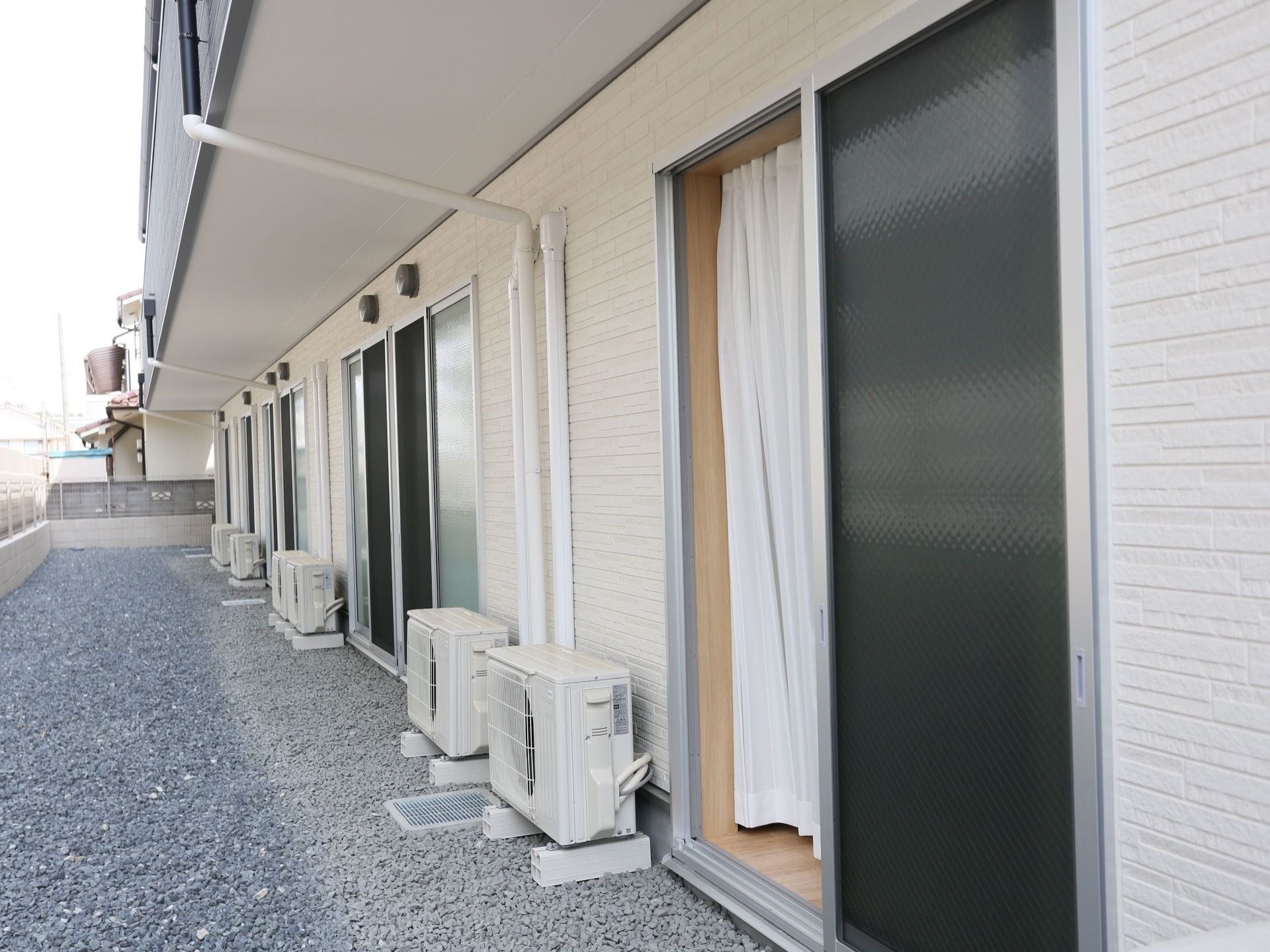 居室のガラス戸から外に出ると、地面に砂利が敷き詰められ、エアコンの外機が並んでいる。前方にフェンスを設置している。