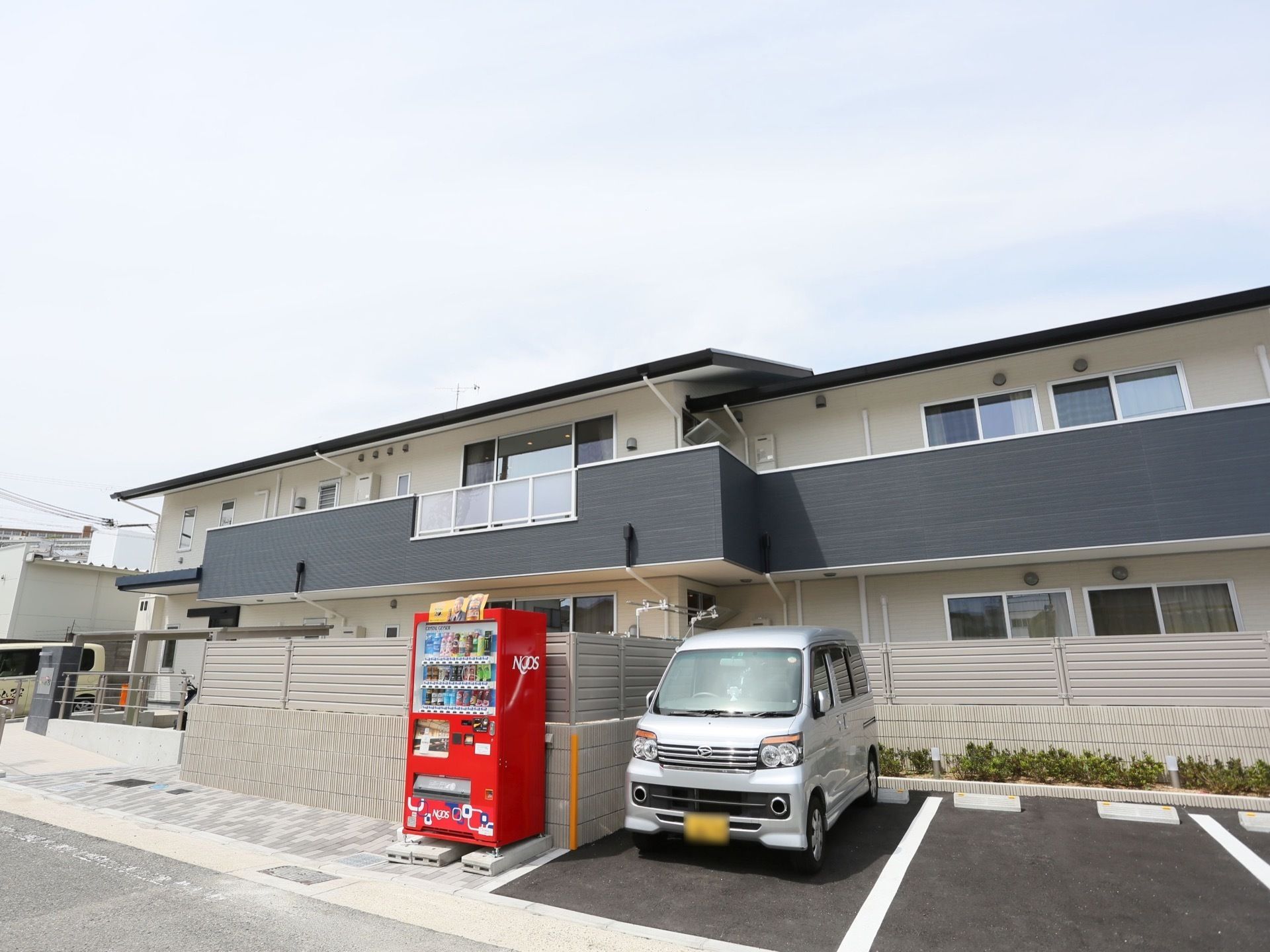 グレー系のツートンカラーの外観である。建物の前方に駐車スペースを設け、フェンス際に自動販売機を設置している。