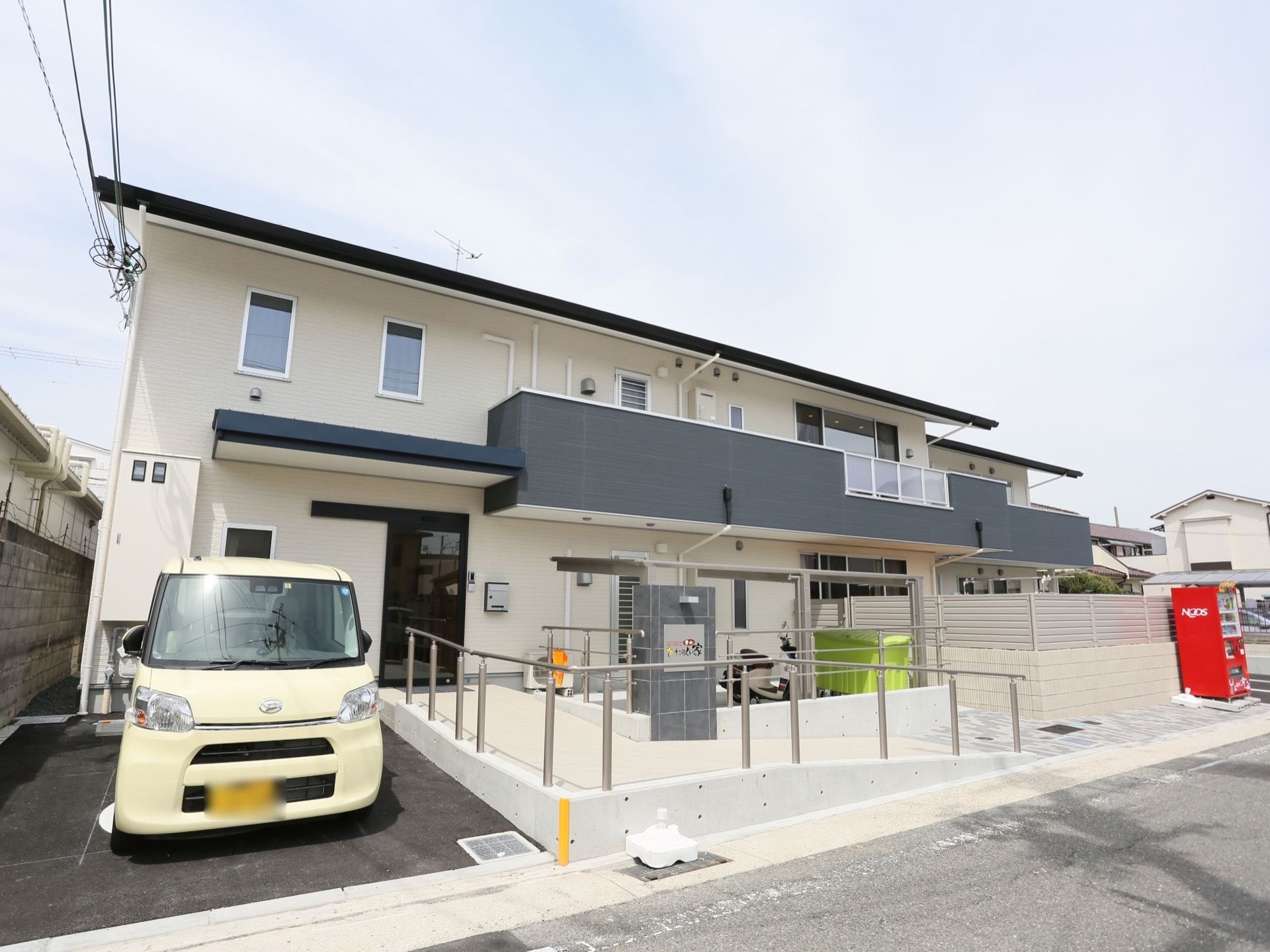 道路に面した自宅のような外観の２階建て建物である。敷地内には、駐車スペースの他に駐輪場も設けている。