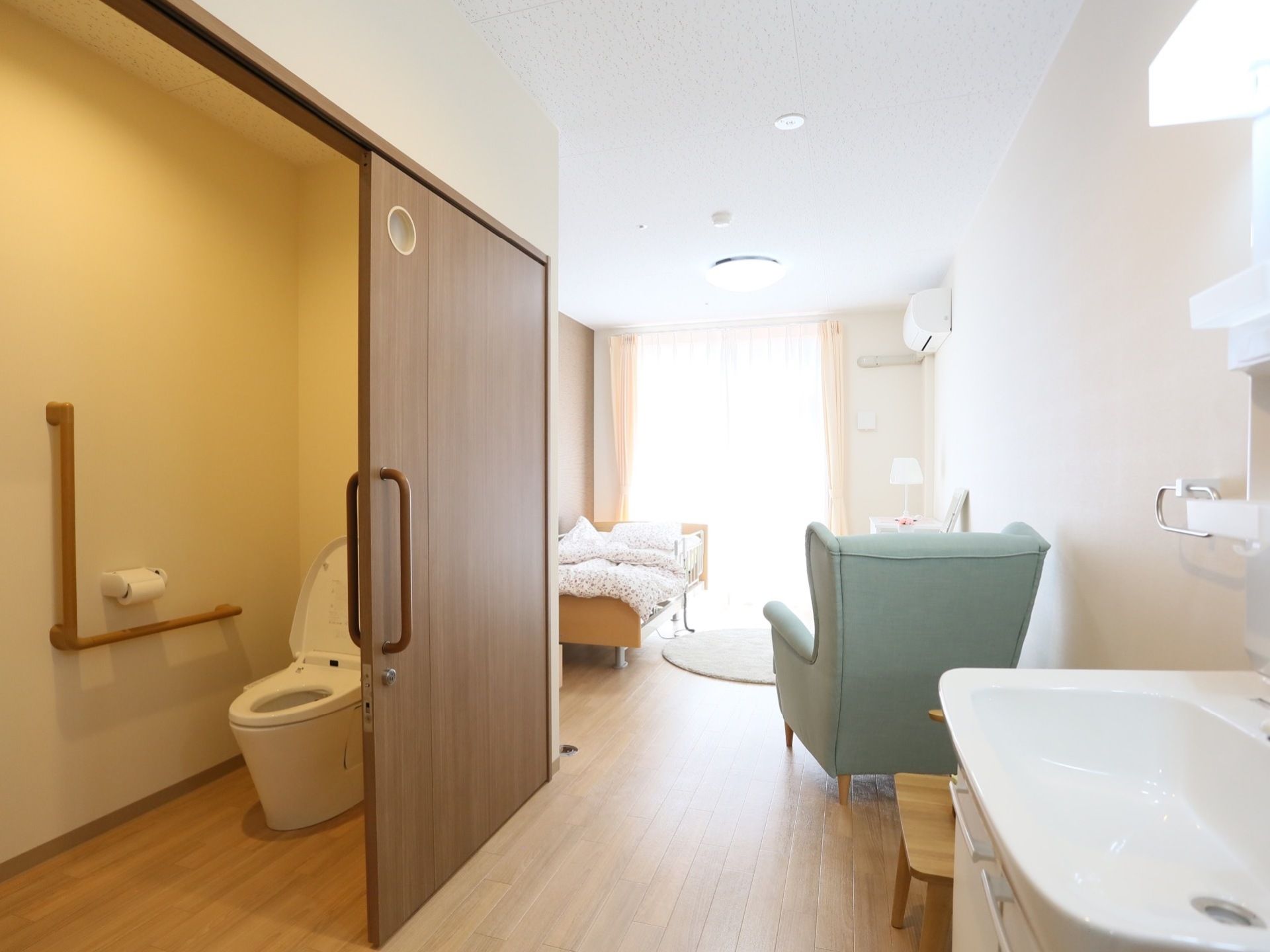 掃出し窓のある居室には、右端に洗面台、左側にトイレがある。茶色の引き戸には、小窓がついている。天井が高く開放感がある。