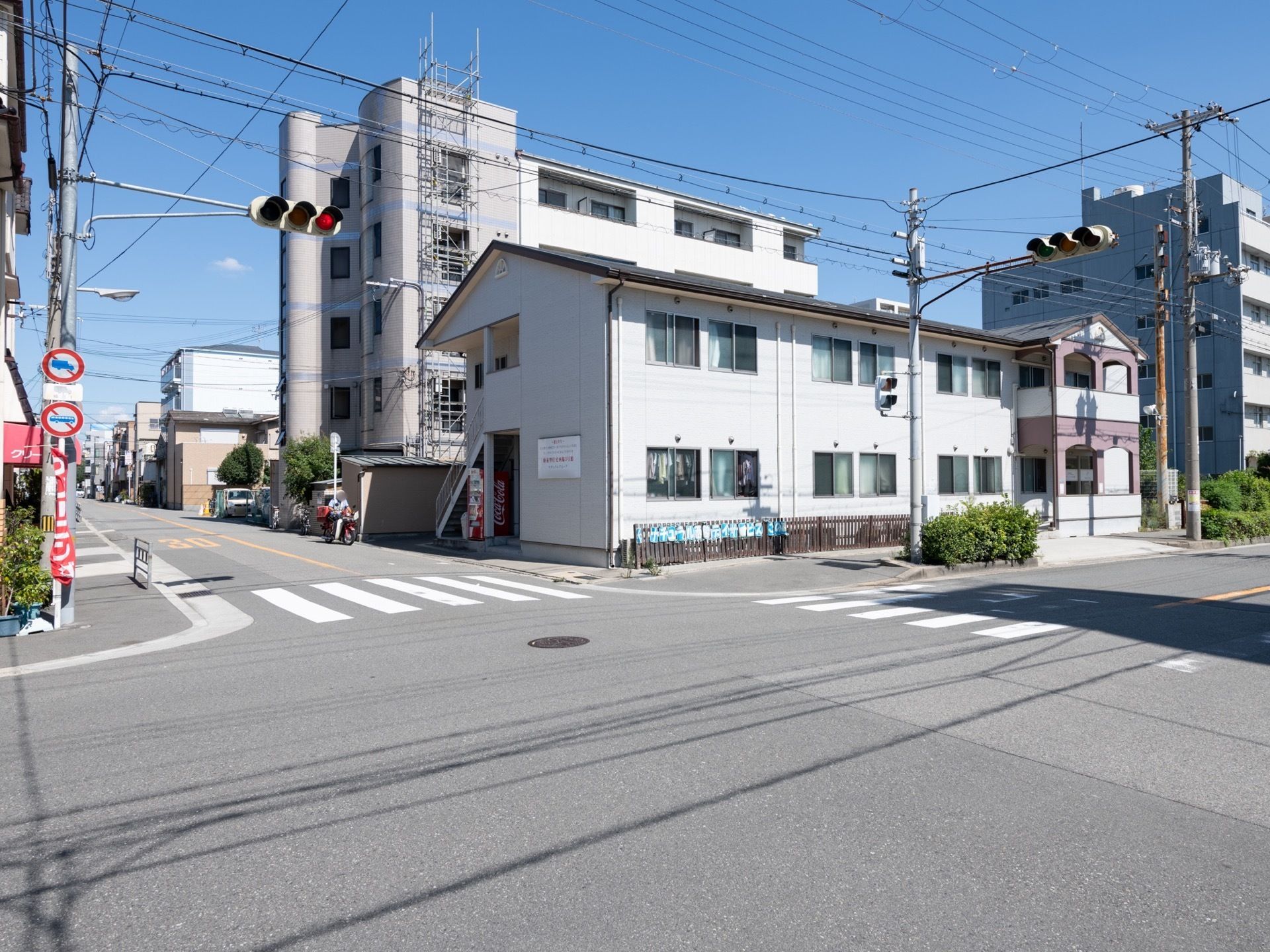 交差点に面した２階建ての建物がある。窓がある壁面には日光が良く当たっている。外階段の下に飲料の自動販売機が設置されている。
