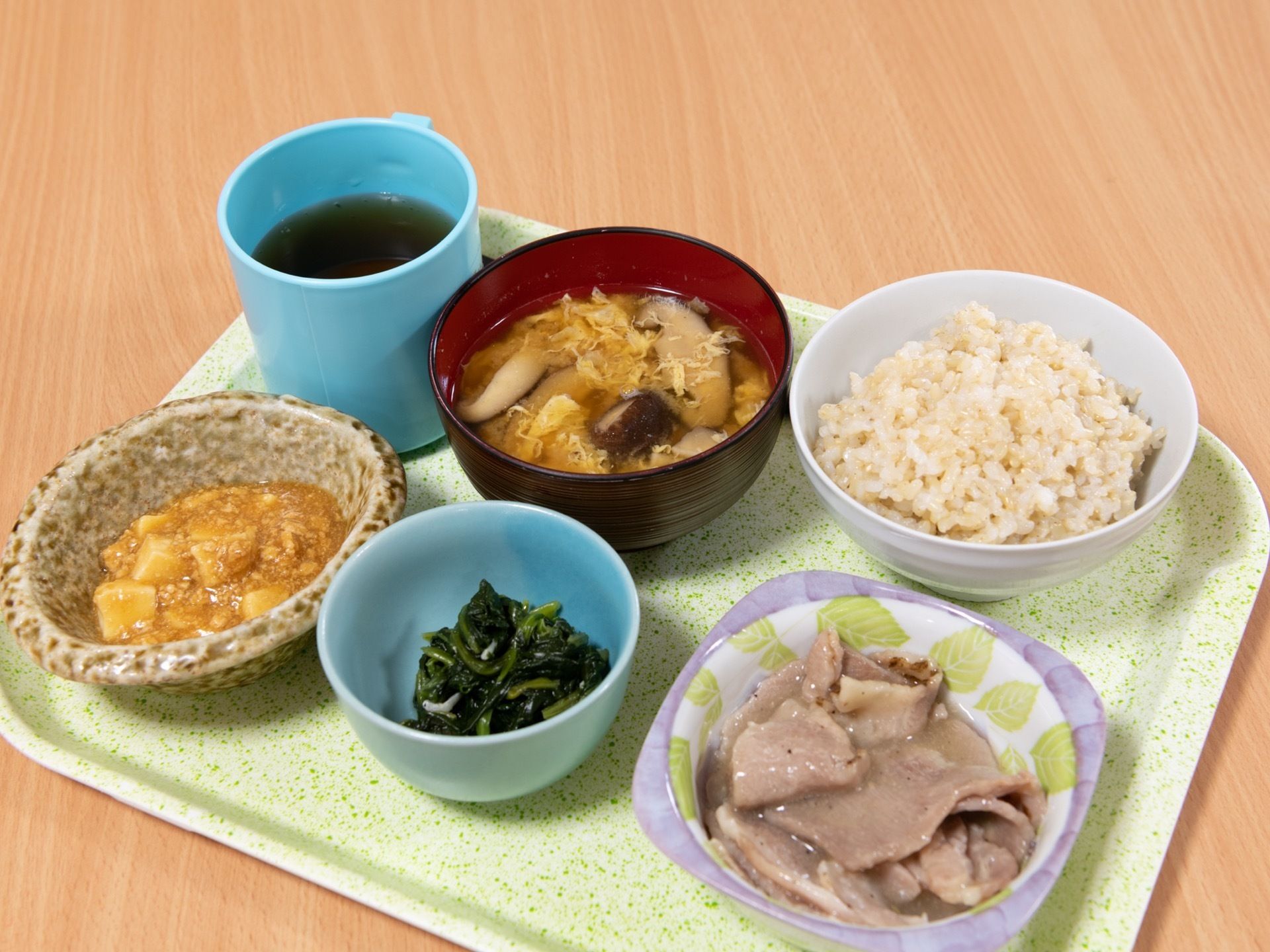 お盆の上にご飯、お味噌汁、お肉料理、お豆腐、おひたしと彩りや栄養バランスの良い献立が並んでいる。コップにはお茶が入っている。