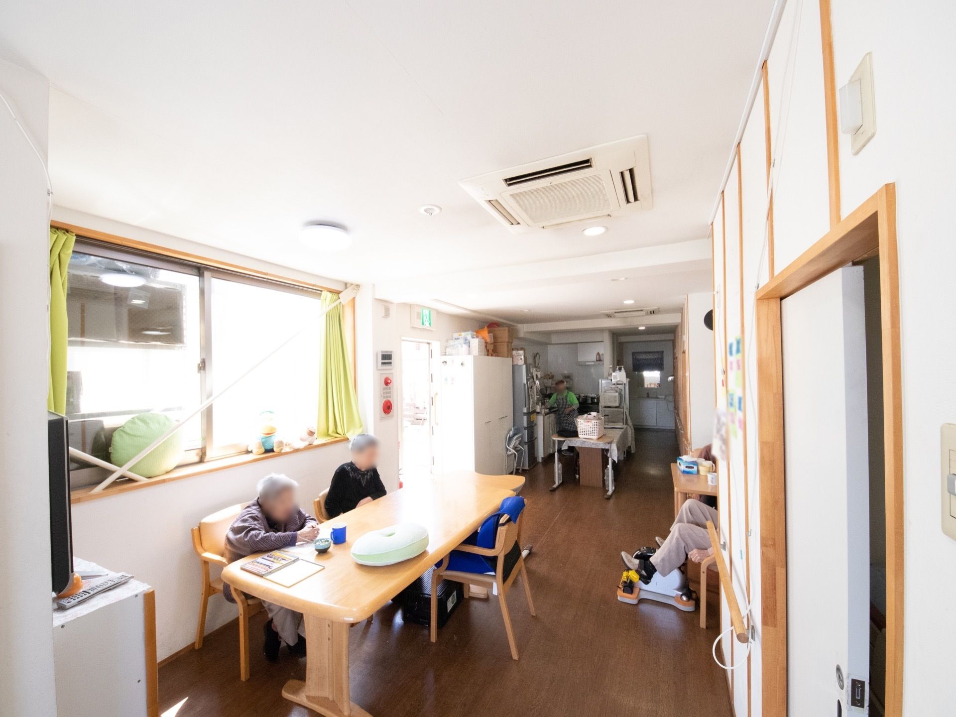 共有スペースは縦長になっている。入居者様が木製のテーブルに座り、お茶を飲んでいる。キッチンが併設されている。