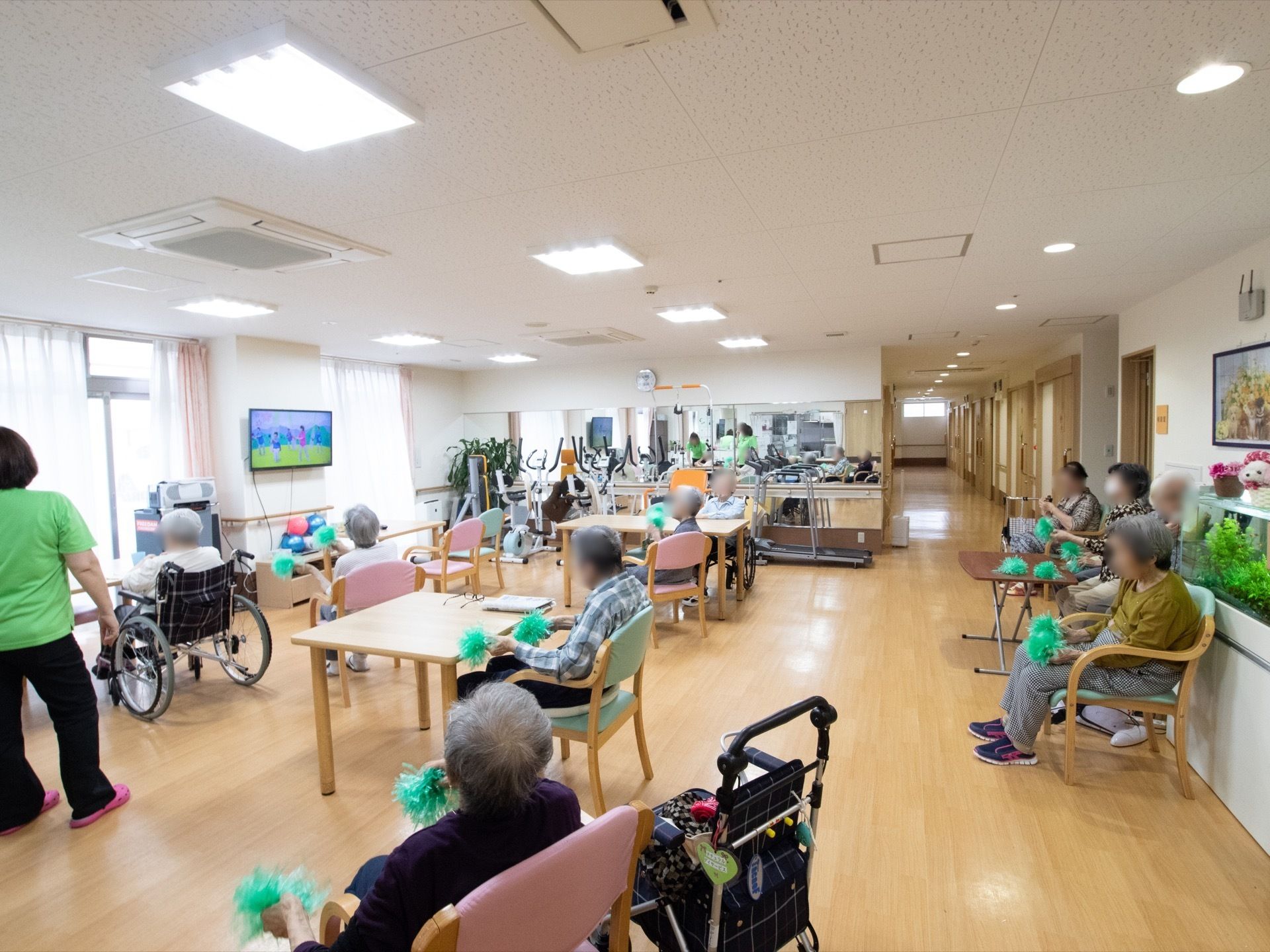 窓の間にある壁にテレビが付いており、入居者様はそちらに向かって座っている。緑のポンポンを持っている。