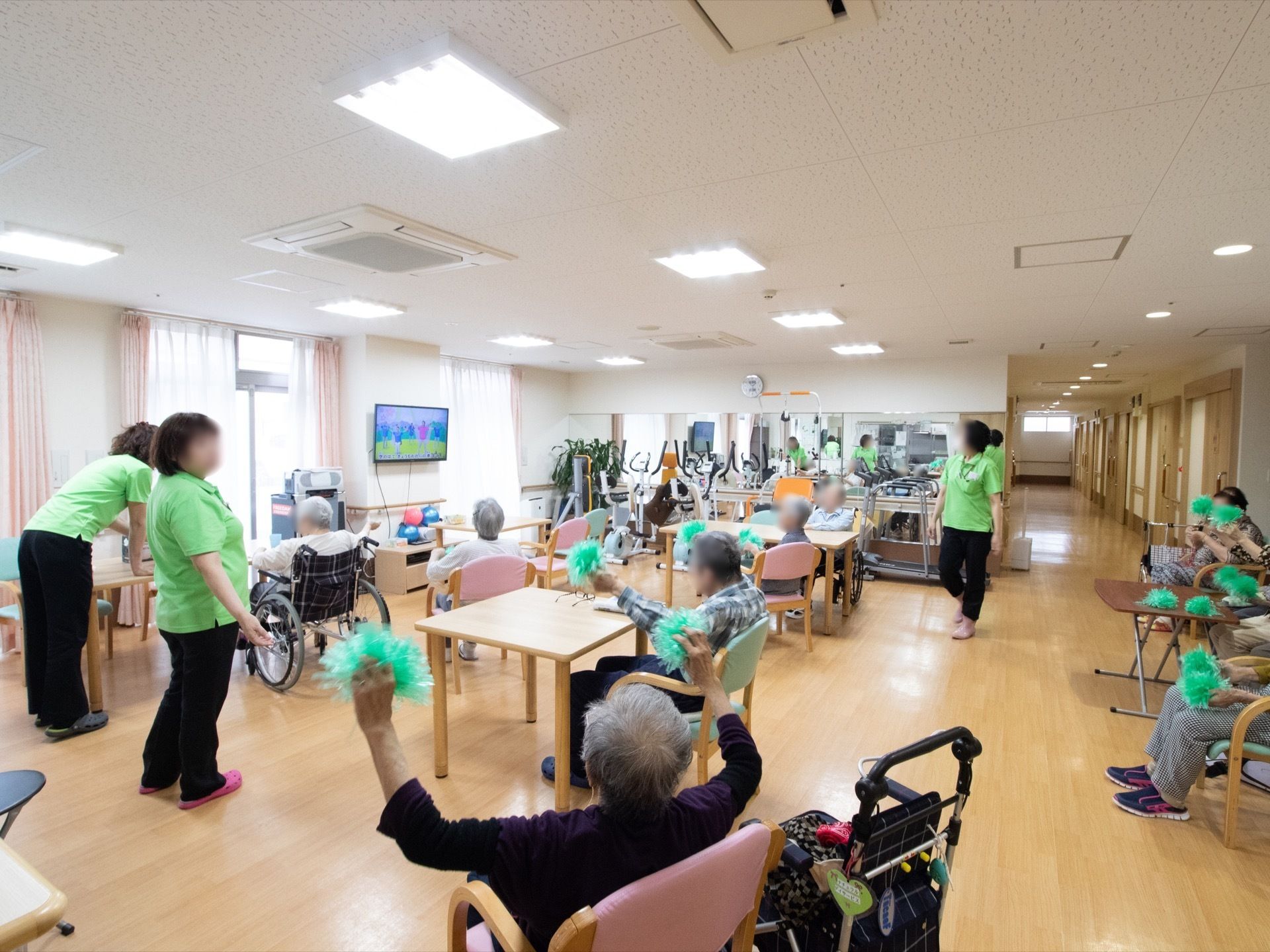 黄緑色のポロシャツを着たスタッフが３人おり、入居者様の間に立っている。リビングの隅には、健康器具が置いてある。