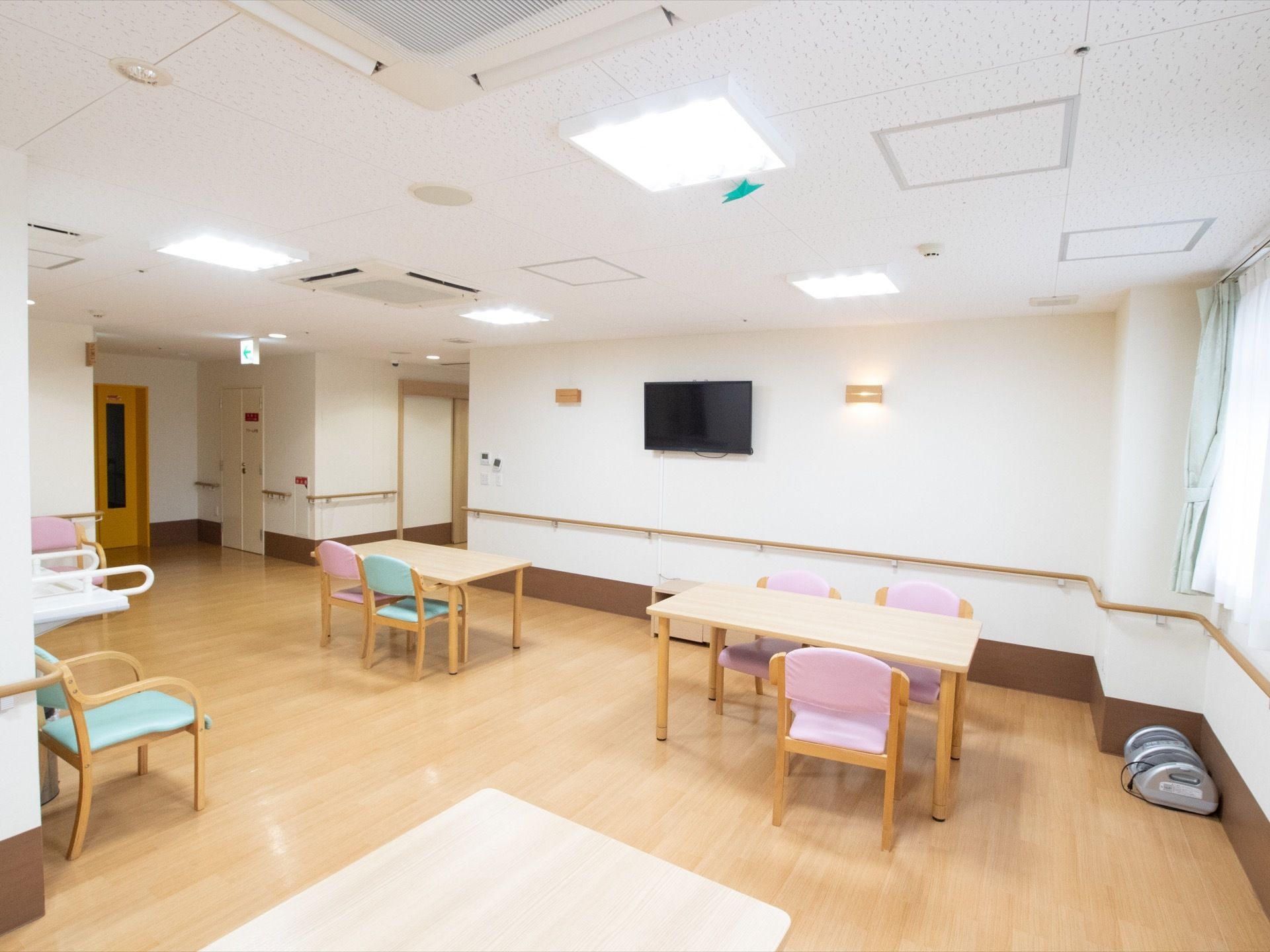 壁にテレビがかけられた、共用スペースである。複数のテーブルセットが設置されている。壁に手すりも完備。