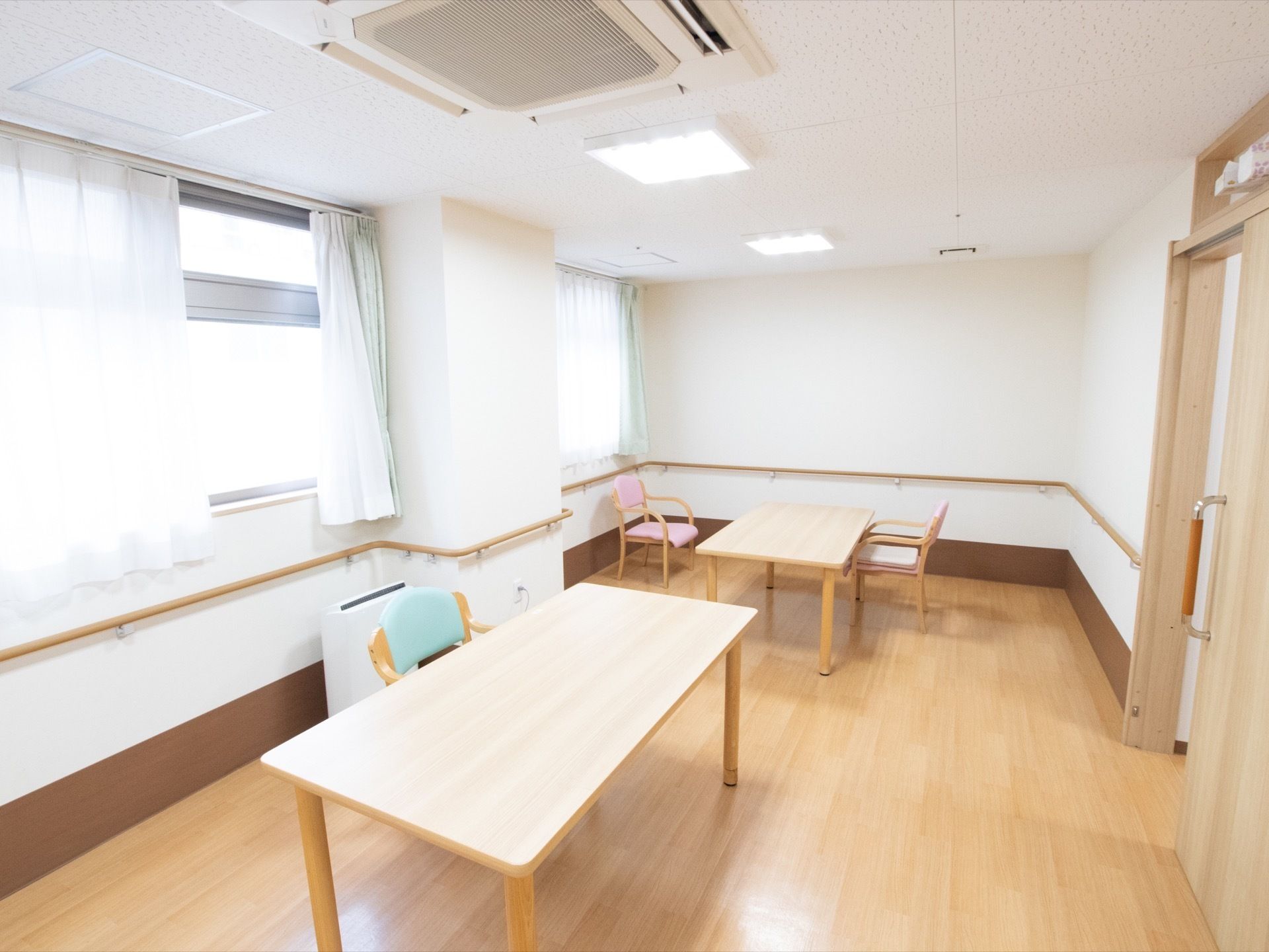 広々したテーブル、グリーンやピンクの椅子が設置されている。大きな窓が備え付けられ、日当たりのよい空間である。