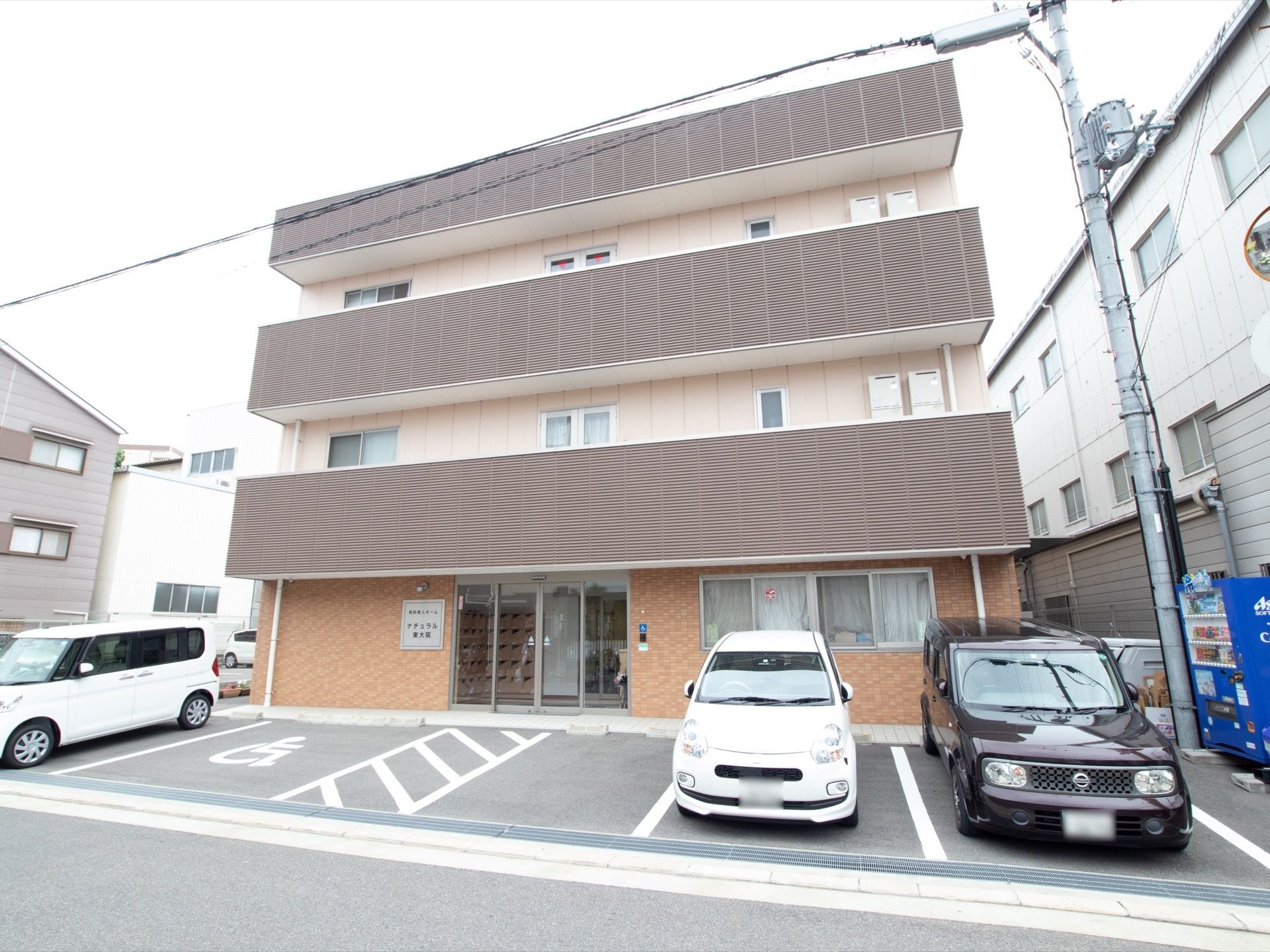 ３階建ての施設でピンクの外壁とオレンジのレンガタイル張りになっている。施設前は駐車スペースになっている。
