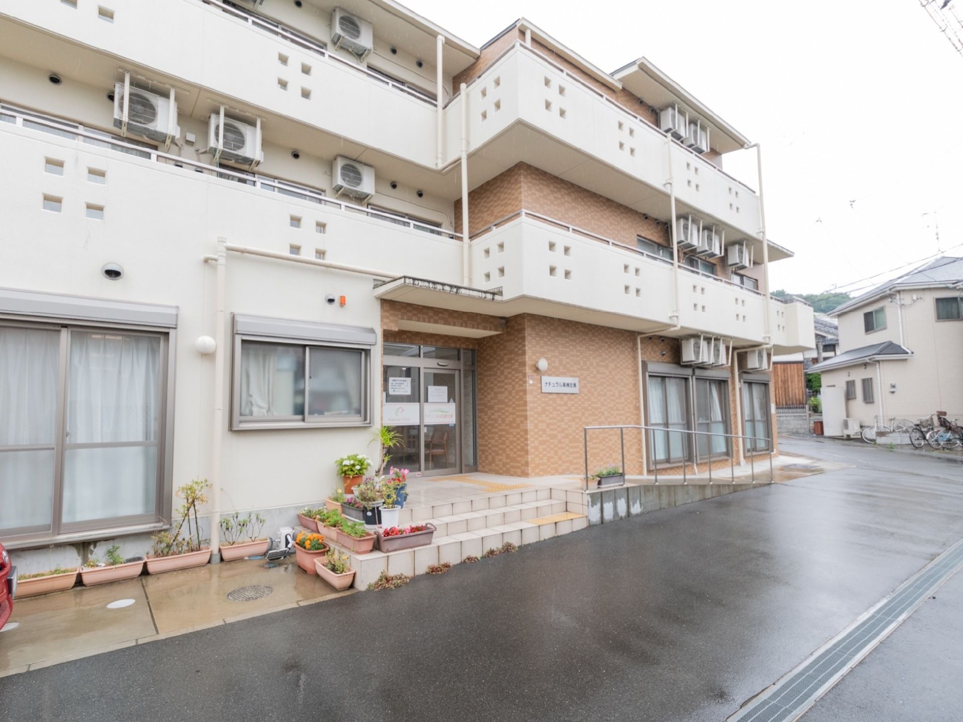 外壁のデザインは、ライトブラウンと白のツートンカラーになっている。玄関の左側には、数多くのプランターや植木鉢が並んでいる。