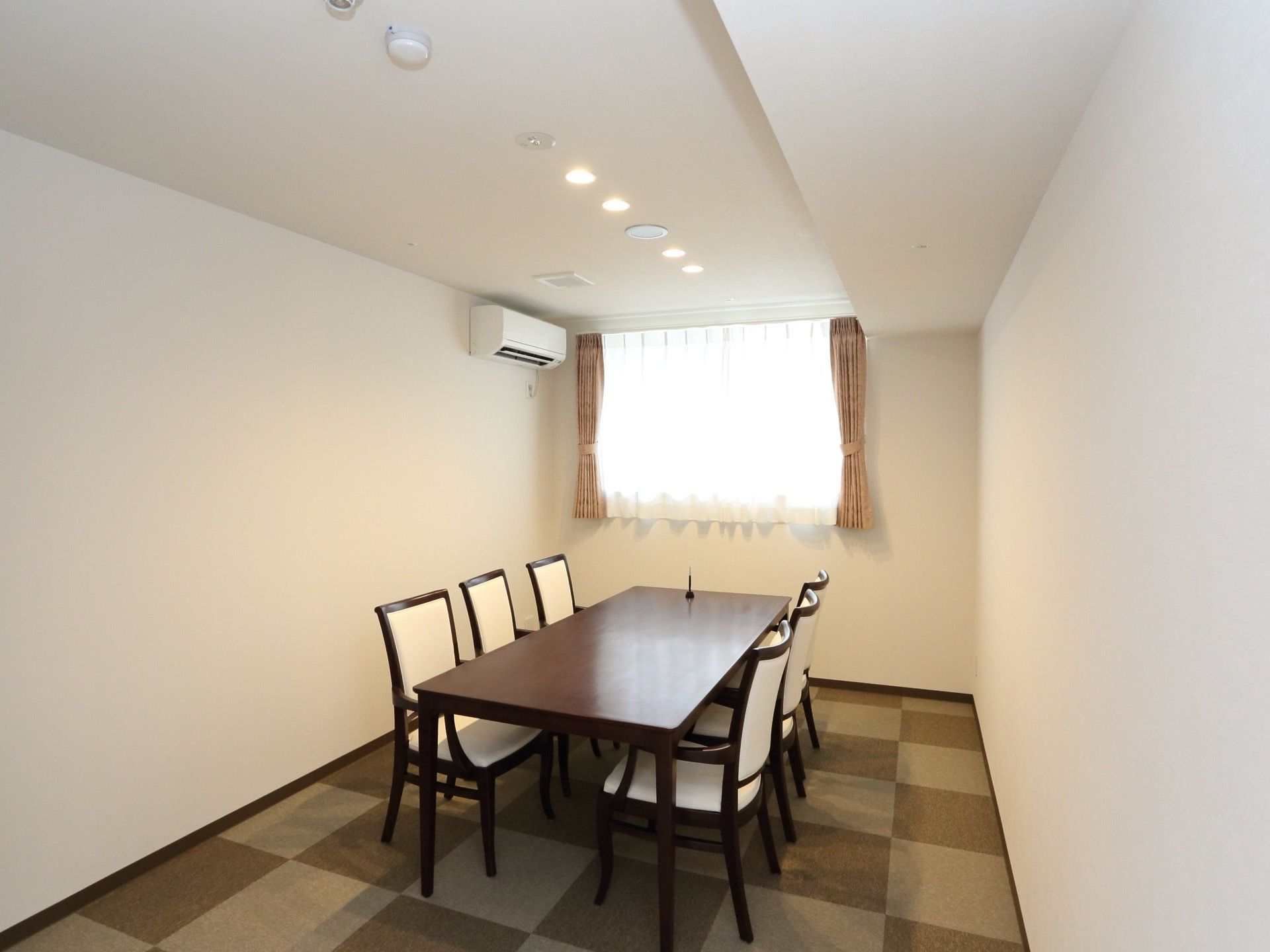 相談室には、部屋の中央に長方形のテーブルと椅子が６脚用意されている。正面にはカーテン付きの窓があり、壁面上部にエアコンが設置されている。