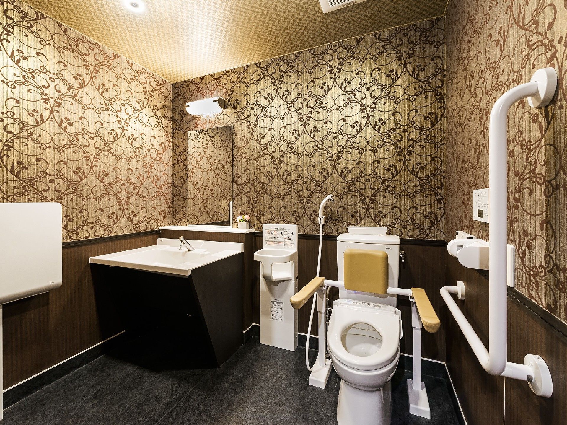 十分な広さがあり、つる草模様の壁紙が落ち着いた雰囲気のトイレである。背もたれ付き両側手すりがあり安定して座ることができる。