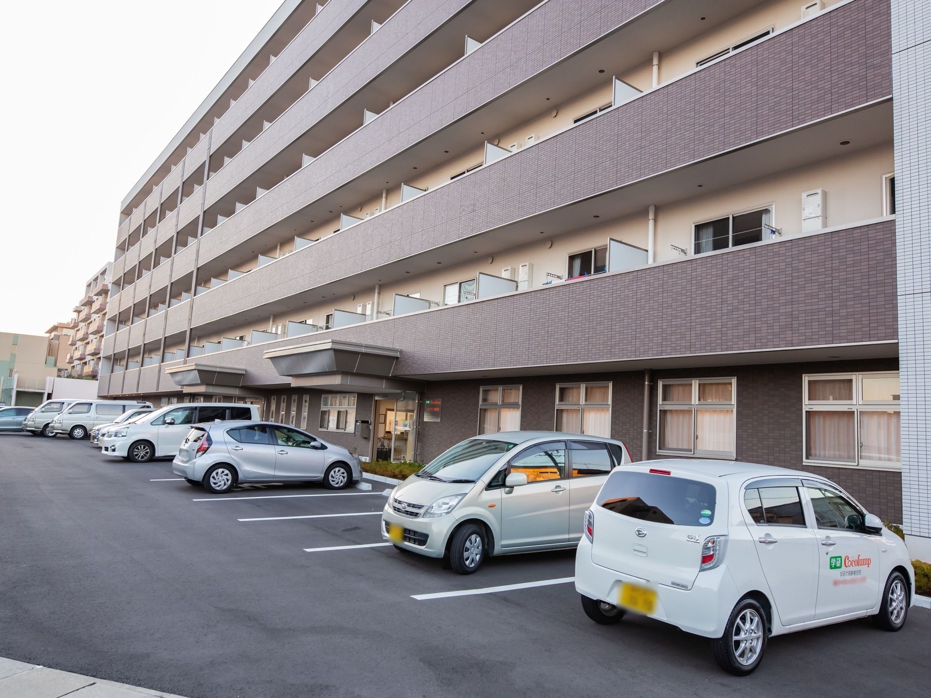 六階建ての建物が見える。建物の前方は駐車場になっており、車が数台駐車している。各部屋のバルコニーが建物正面側にある。