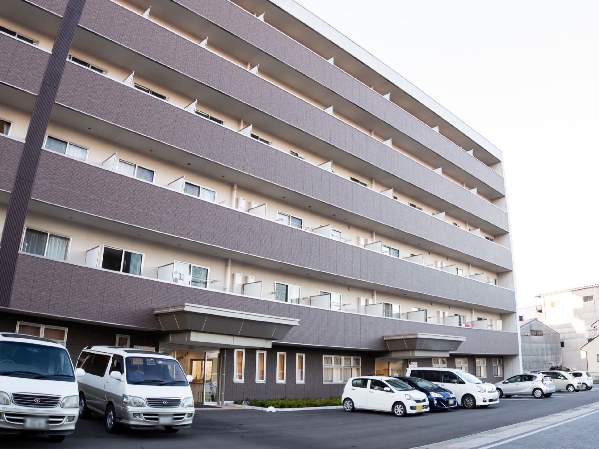 施設は6階建てでダークブラウンの外観である。建物の前には駐車スペースがあり、複数の車が停められている。