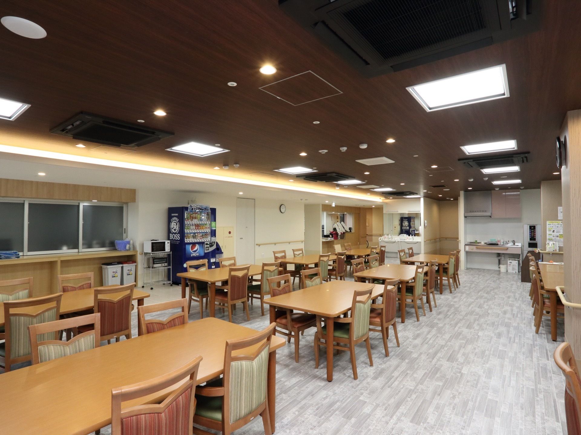 天井はシックな茶色、床は明るいグレーで施された食堂である。複数のダイニングテーブル、椅子が規則的に配置されている。
