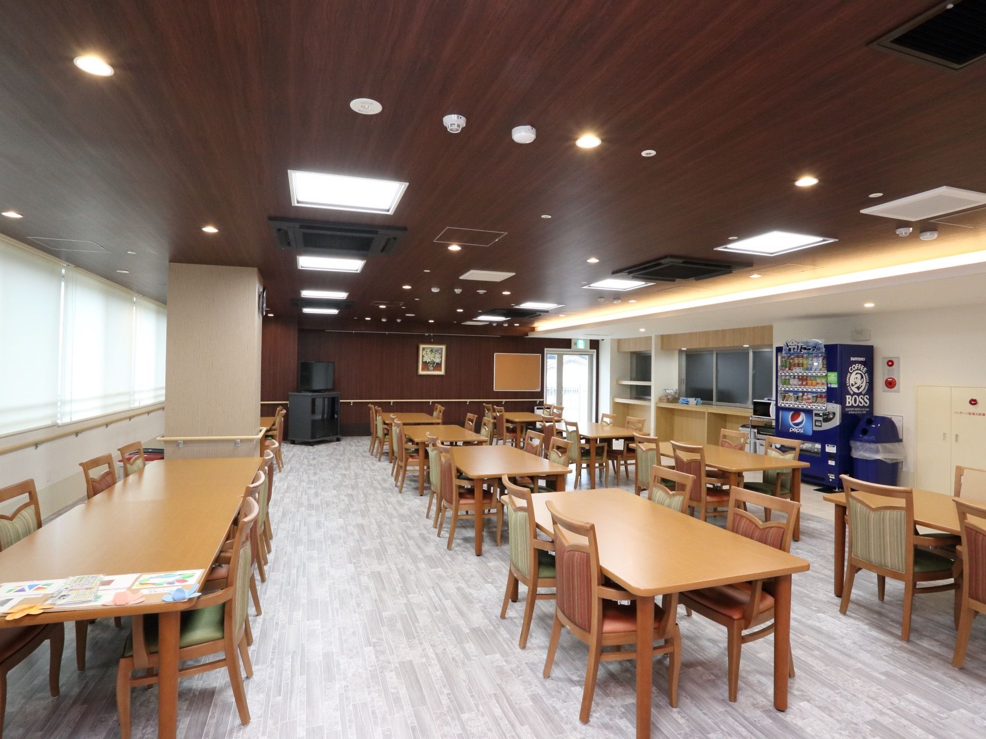 天井に丸型、四角形の照明が複数設置されている。広々とした木製のテーブルに、背もたれ椅子が置かれてある。
