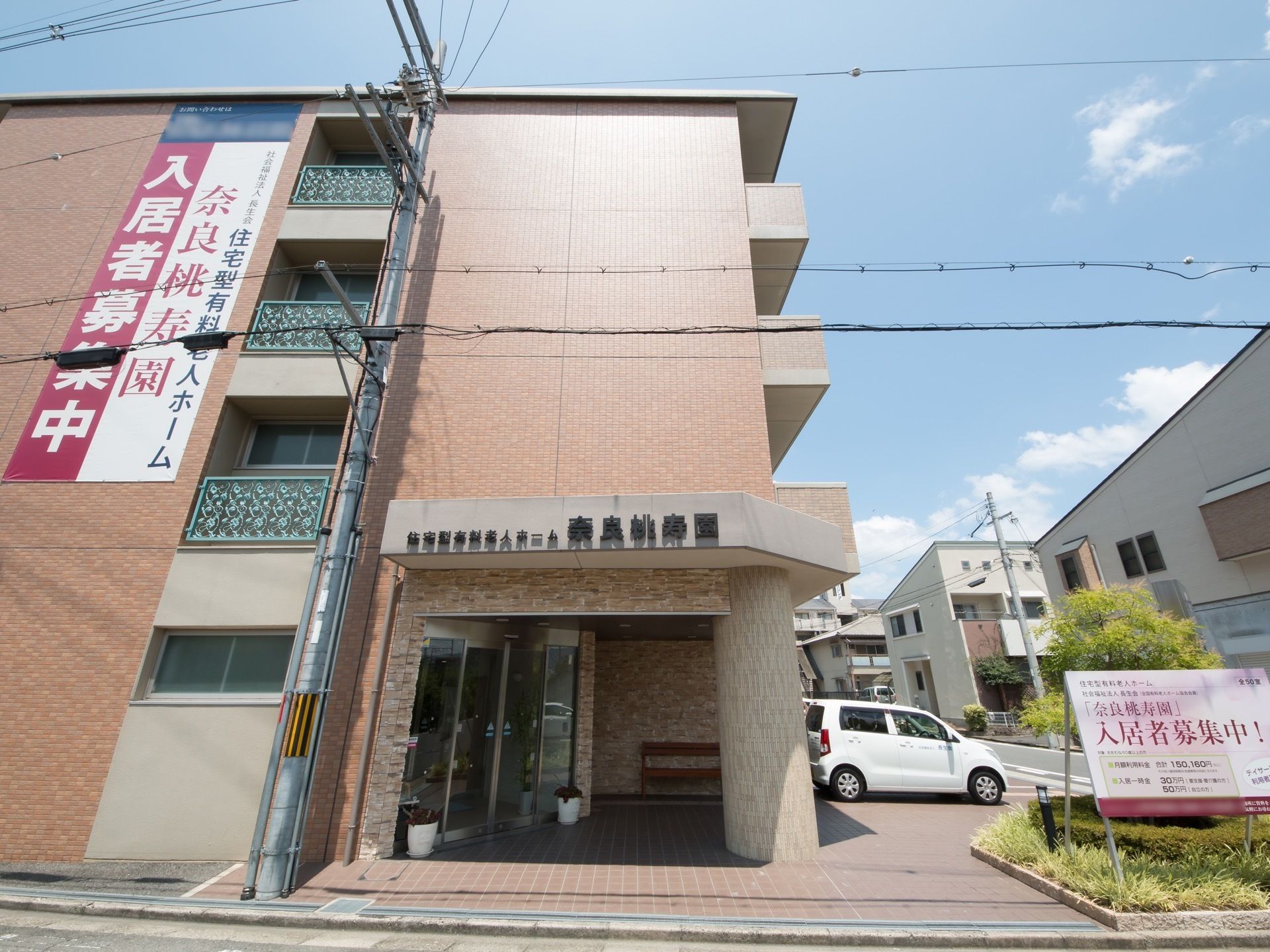 ４階建てのレンガ調の建物に入居者募集中の横断幕が掛けられている。茶色いタイルが敷かれた駐車場に白い車が停められている。