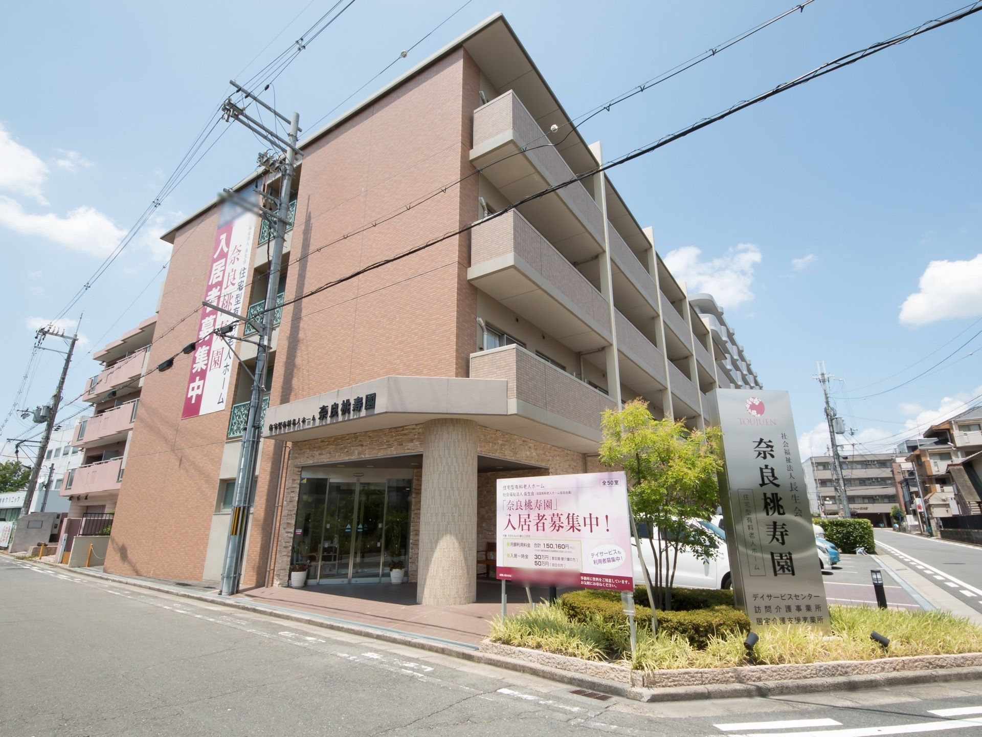 敷地内の植栽スペースに樹木が植えられ、施設の名称が表示された看板と、入居者募集中の看板が立っている。