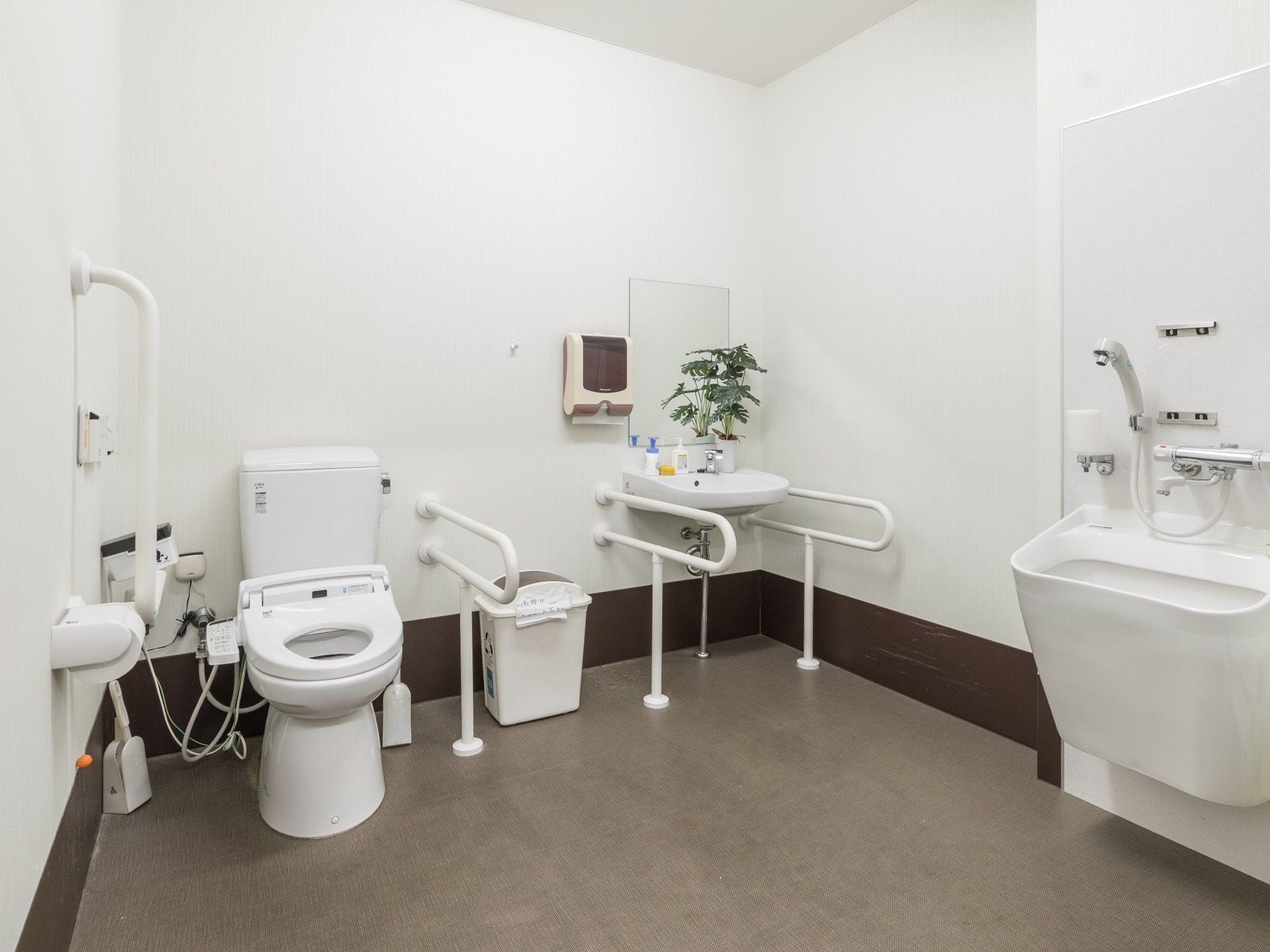 白い壁とブラウンの床のトイレ。トイレ内には汚物用の流し台が設置されている。壁には手すりと緊急通報装置が取り付けられている。