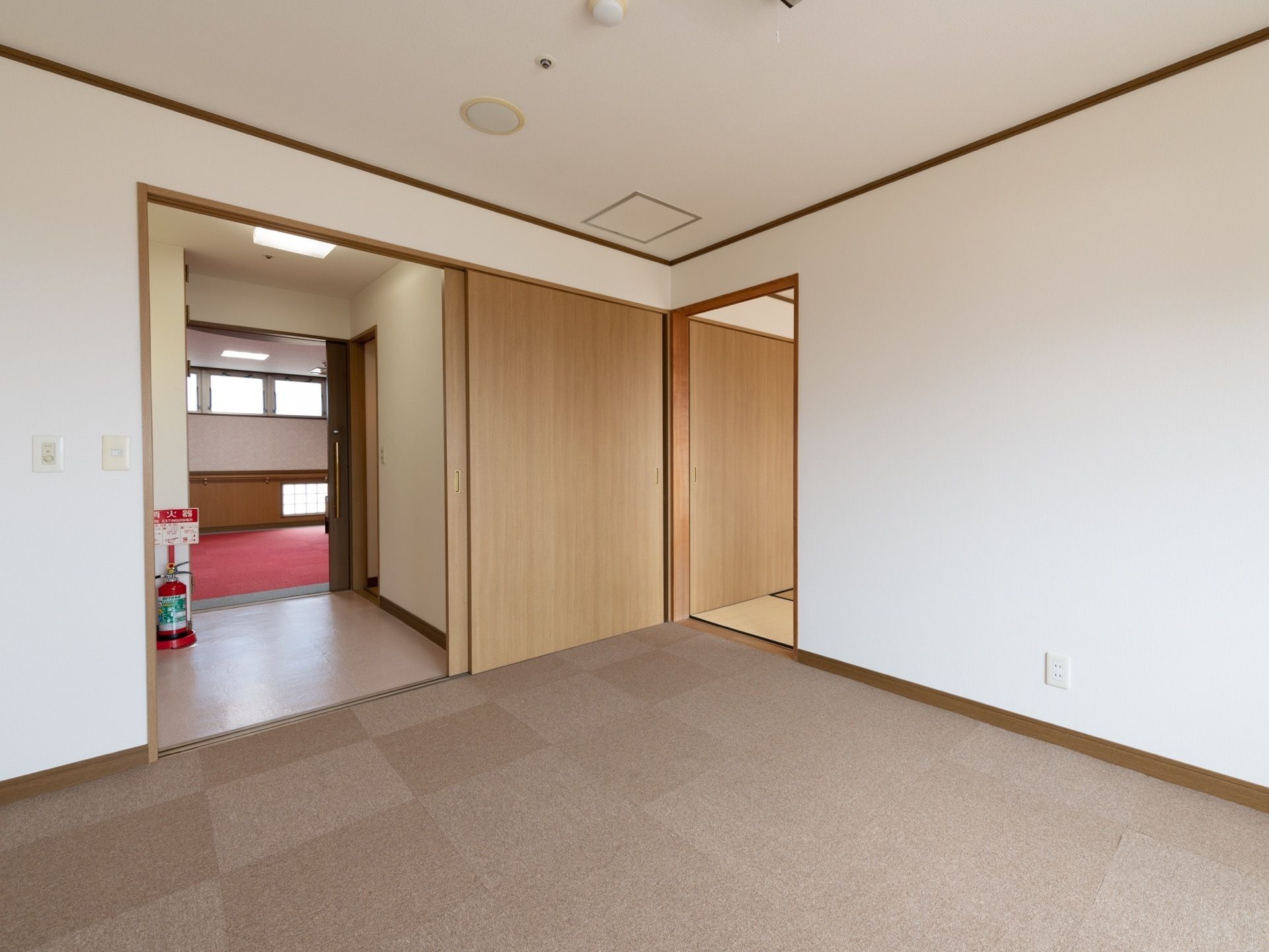 絨毯の広い部屋と、隣に畳の部屋がある入居者様の居室。部屋は扉で仕切られており、消火器が置かれている。