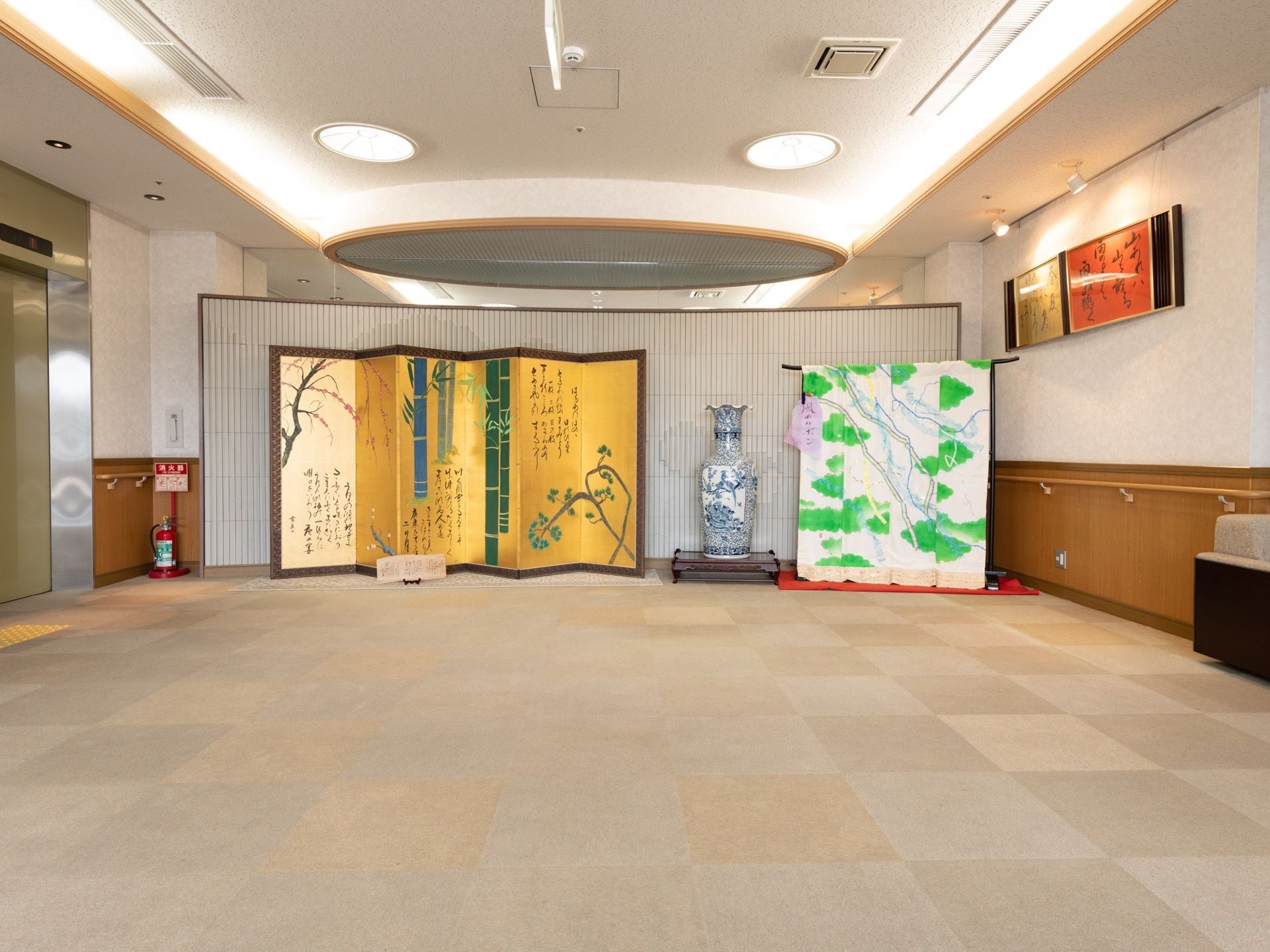 金の屏風と緑色と白の着物、大きな壺が飾られている広いホールがある。壁には書が飾られ、手すりも設置されている。
